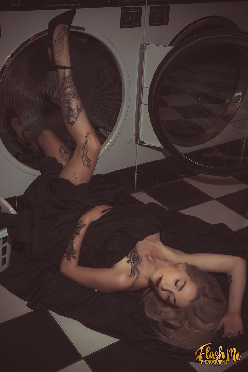 Tattooed woman in a black dress lying dramatically on a laundromat floor with legs in a dryer.
