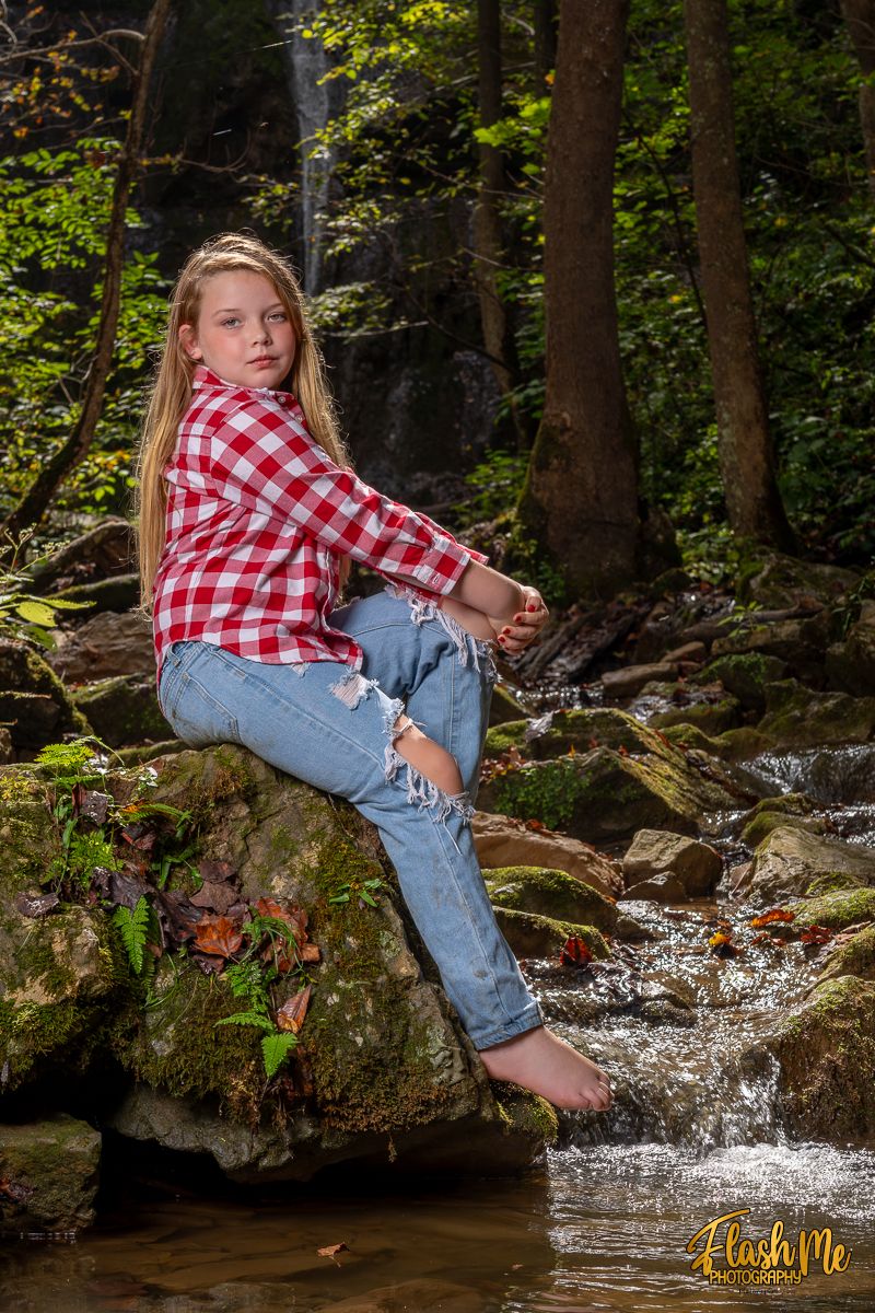 Young girl in red plaid shirt and ripped jeans sitting barefoot on rocks beside a forest stream.