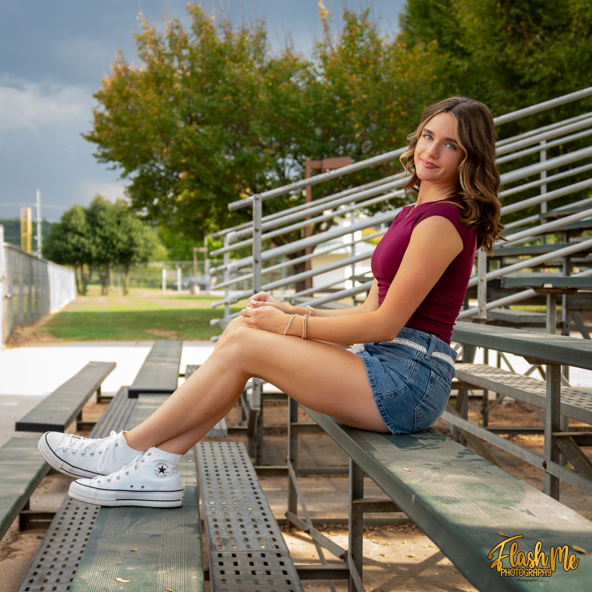 Young woman in maroon top and denim shorts posing on bleachers at an outdoor sports field on a cloudy day.