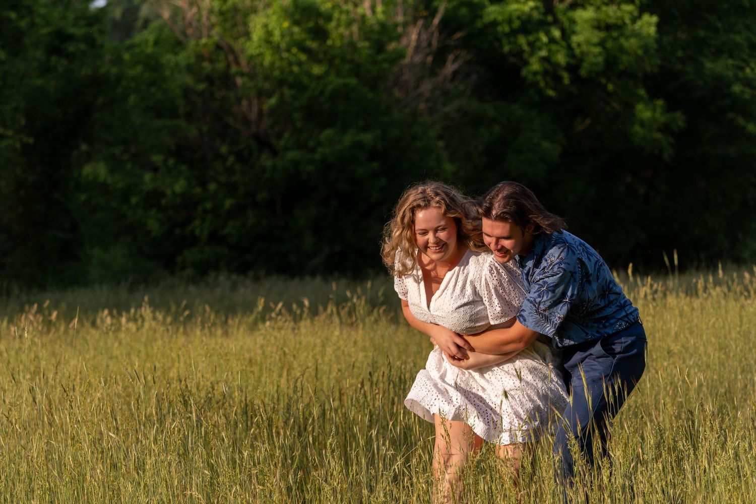 Couple playfully embracing and laughing together in a beautiful green meadow at sunset.