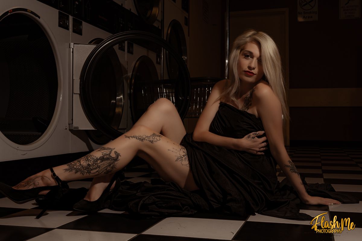 Blonde woman with leg tattoos in a black dress posing on a checkered floor in a laundromat.