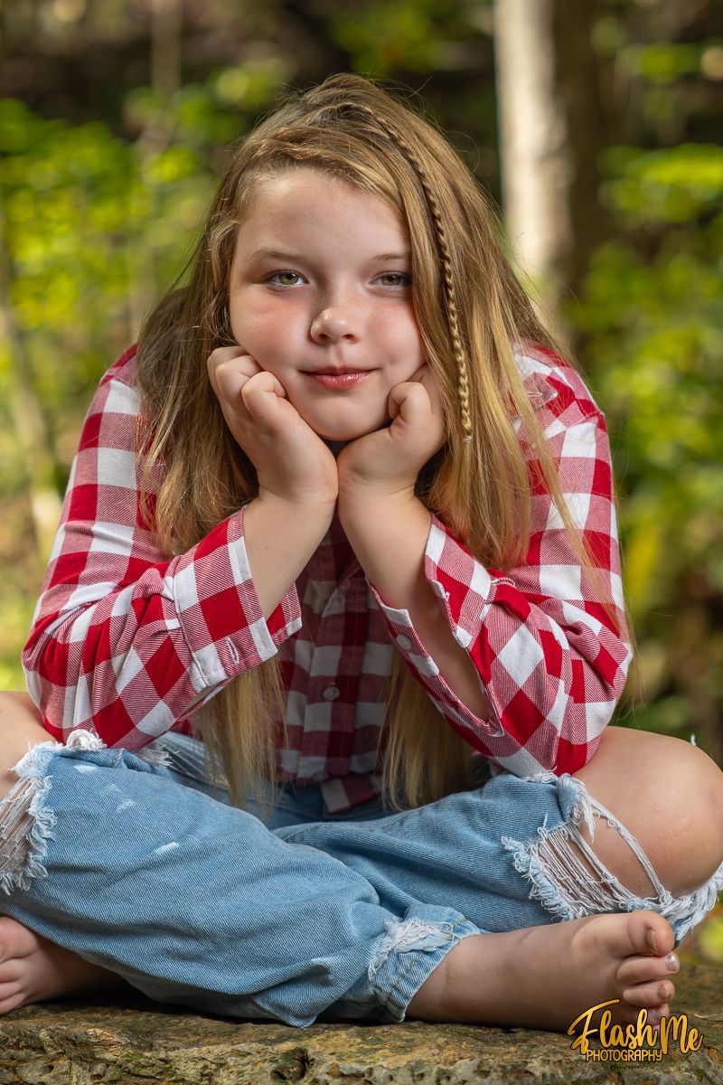Young girl in red plaid shirt and ripped jeans sitting cross-legged outdoors with chin resting on hands.