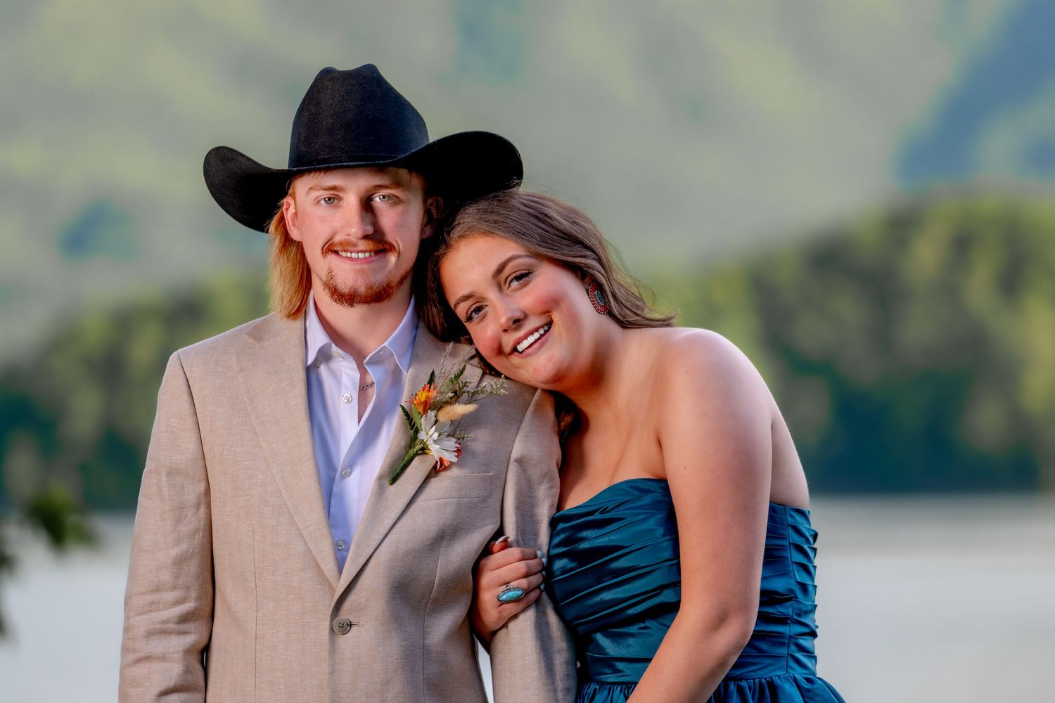 Smiling prom couple posing together outdoors with mountains and lake in the background.