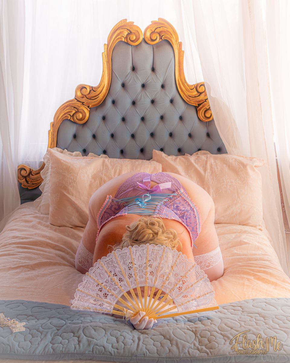 Woman in purple corset lying face down on bed holding a white lace fan.