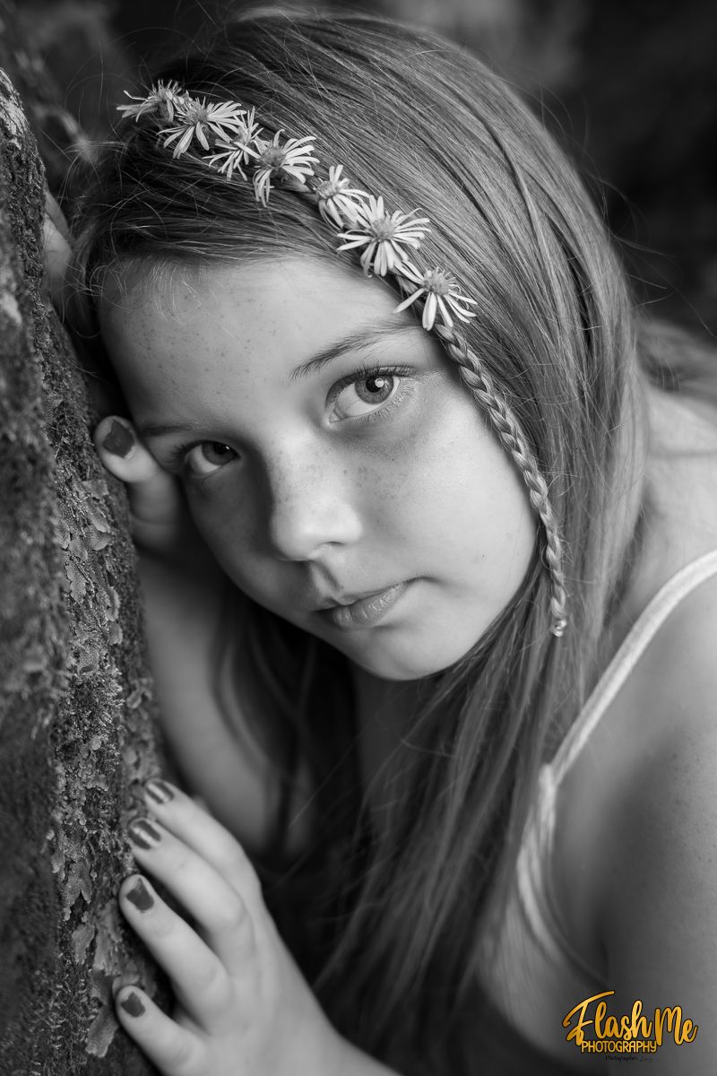 Black and white portrait of a young girl with floral headband leaning against a tree in nature.