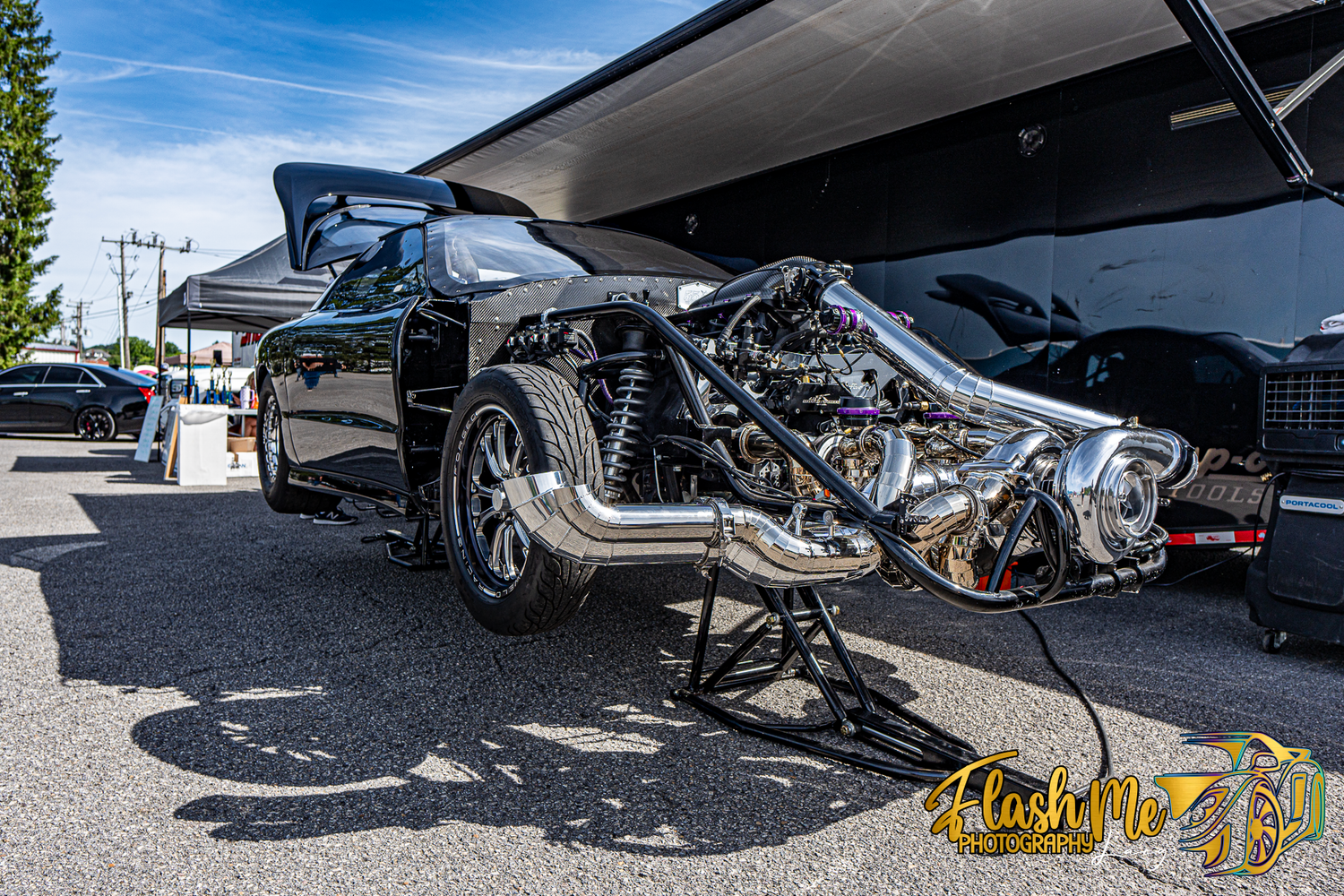 Custom black hot rod car with exposed chrome supercharged engine on display at an outdoor car show.