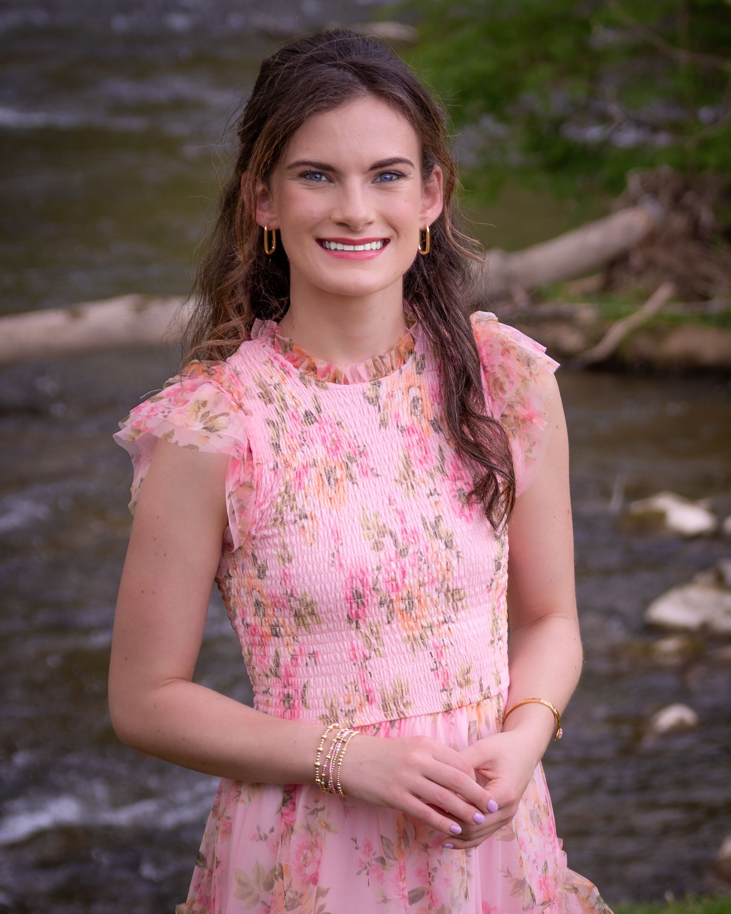 Smiling young woman in a pink floral dress posing near a stream with rocks and trees in the background.