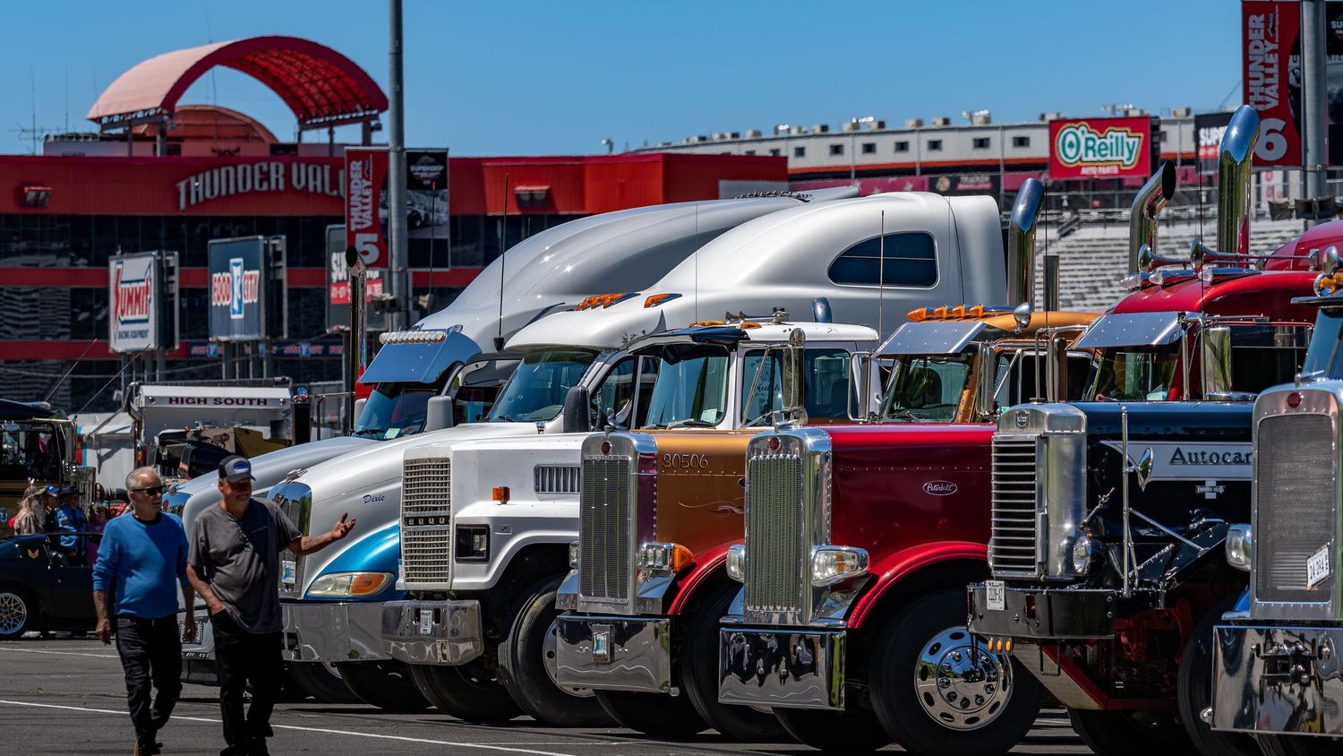 Row of colorful Peterbilt and Kenworth semi trucks parked at a large outdoor truck show.