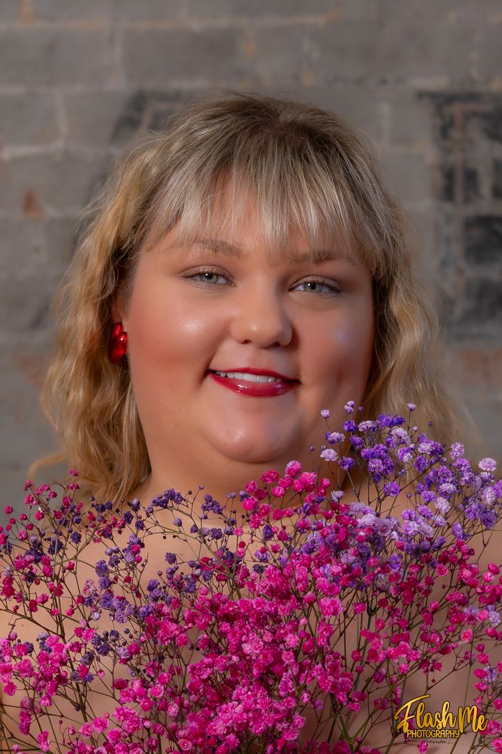 Smiling young woman with blonde hair holding vibrant pink and purple flowers against a brick wall backdrop.