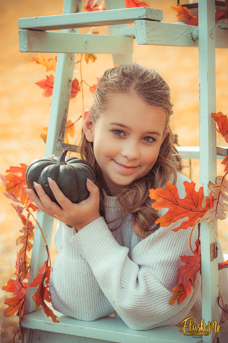 A smiling young girl in a white sweater holds a small pumpkin framed by autumn leaves on a ladder.