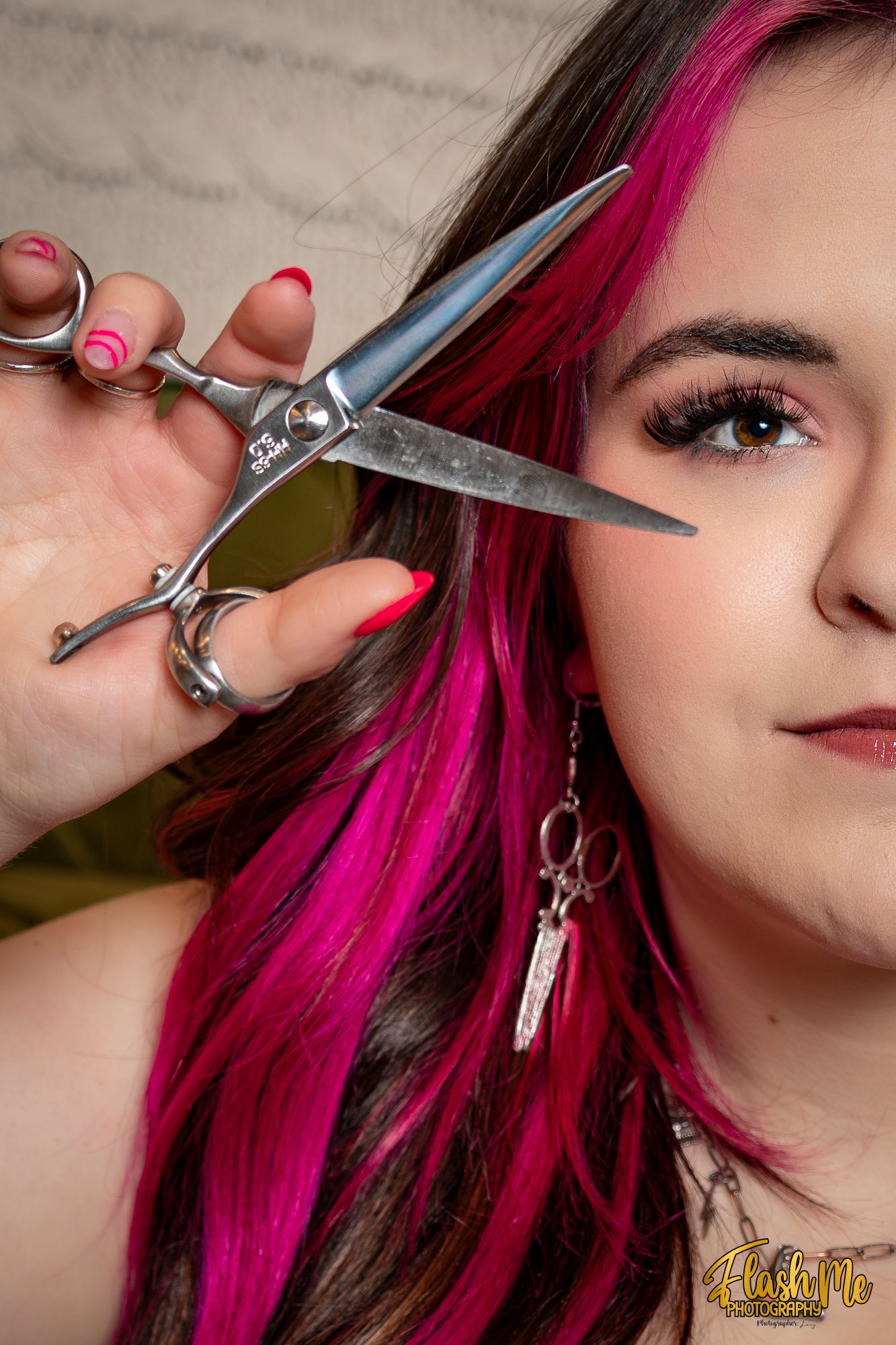 Close-up portrait of a woman with pink-highlighted hair holding scissors near her face with red nails.
