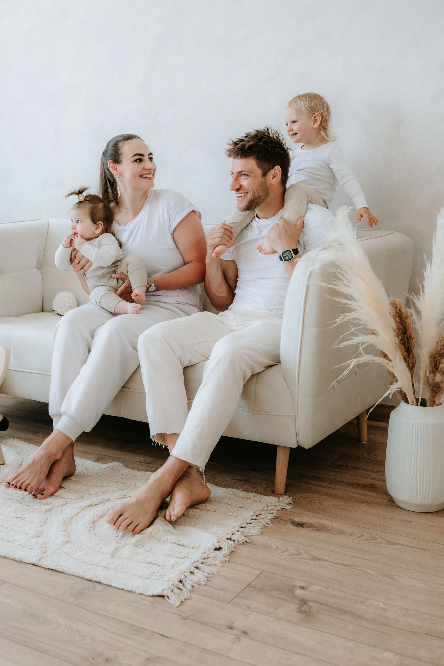 Happy young family with two toddlers sitting together on a white sofa in a bright, modern living room.