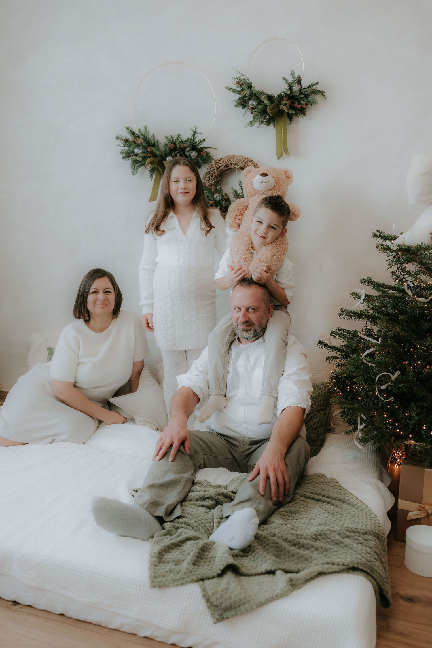 Family of four poses in white outfits near decorated Christmas tree with festive wreaths.
