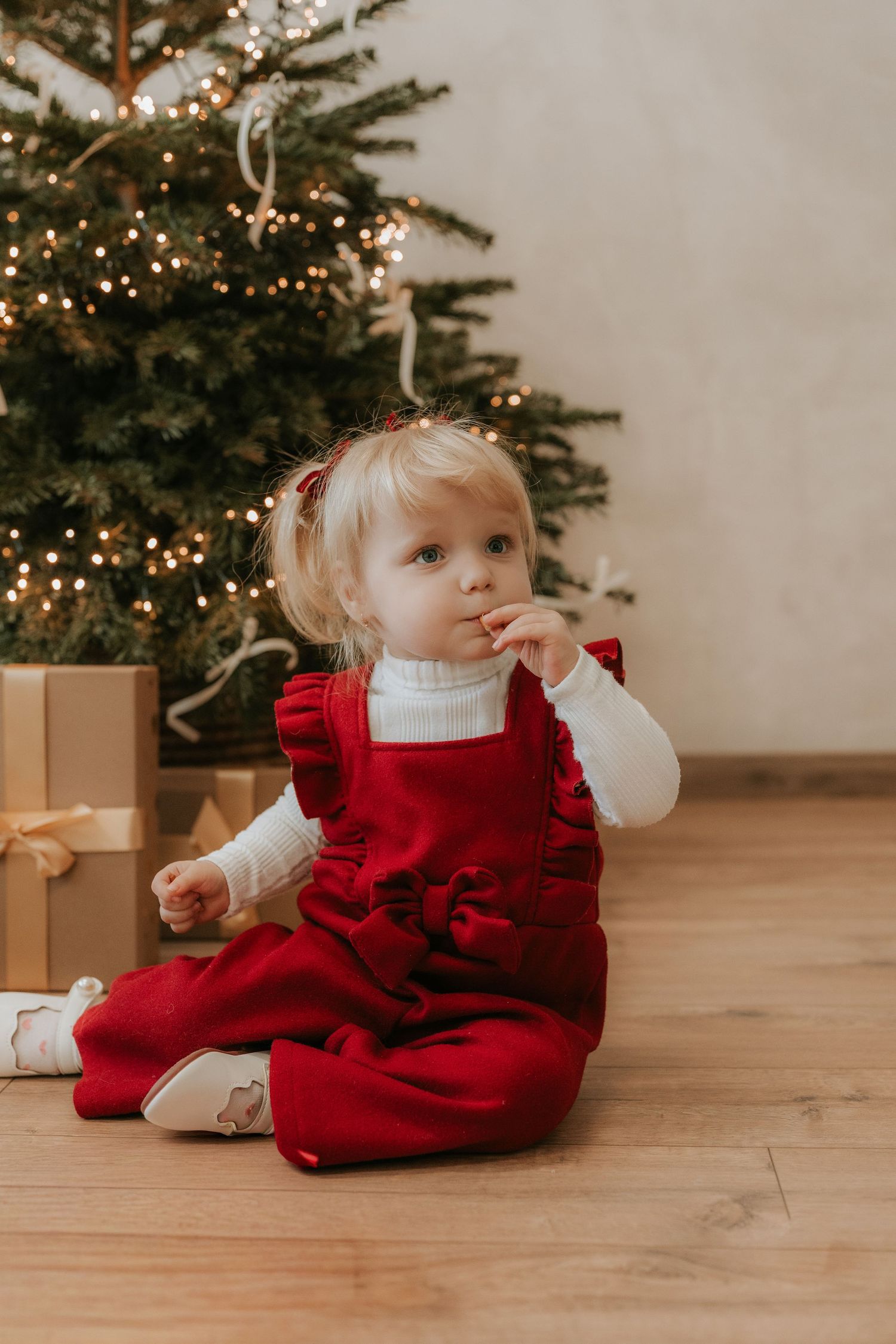 Toddler girl in red ruffle dress sits on wood floor by decorated Christmas tree with gifts.