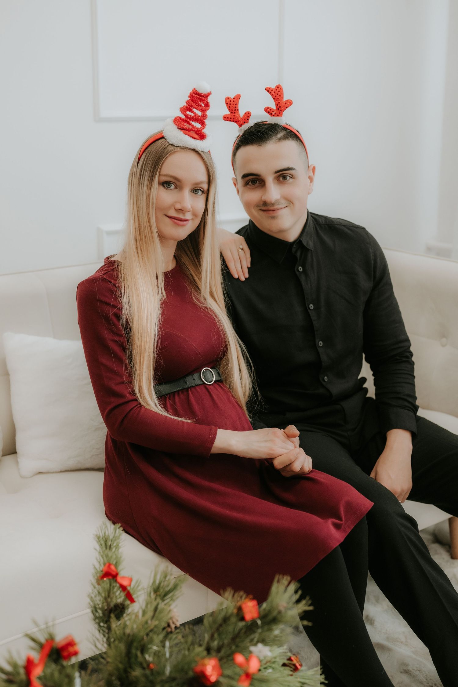 Couple wearing reindeer antler headbands sitting on a white sofa with Christmas decorations for a holiday photo.