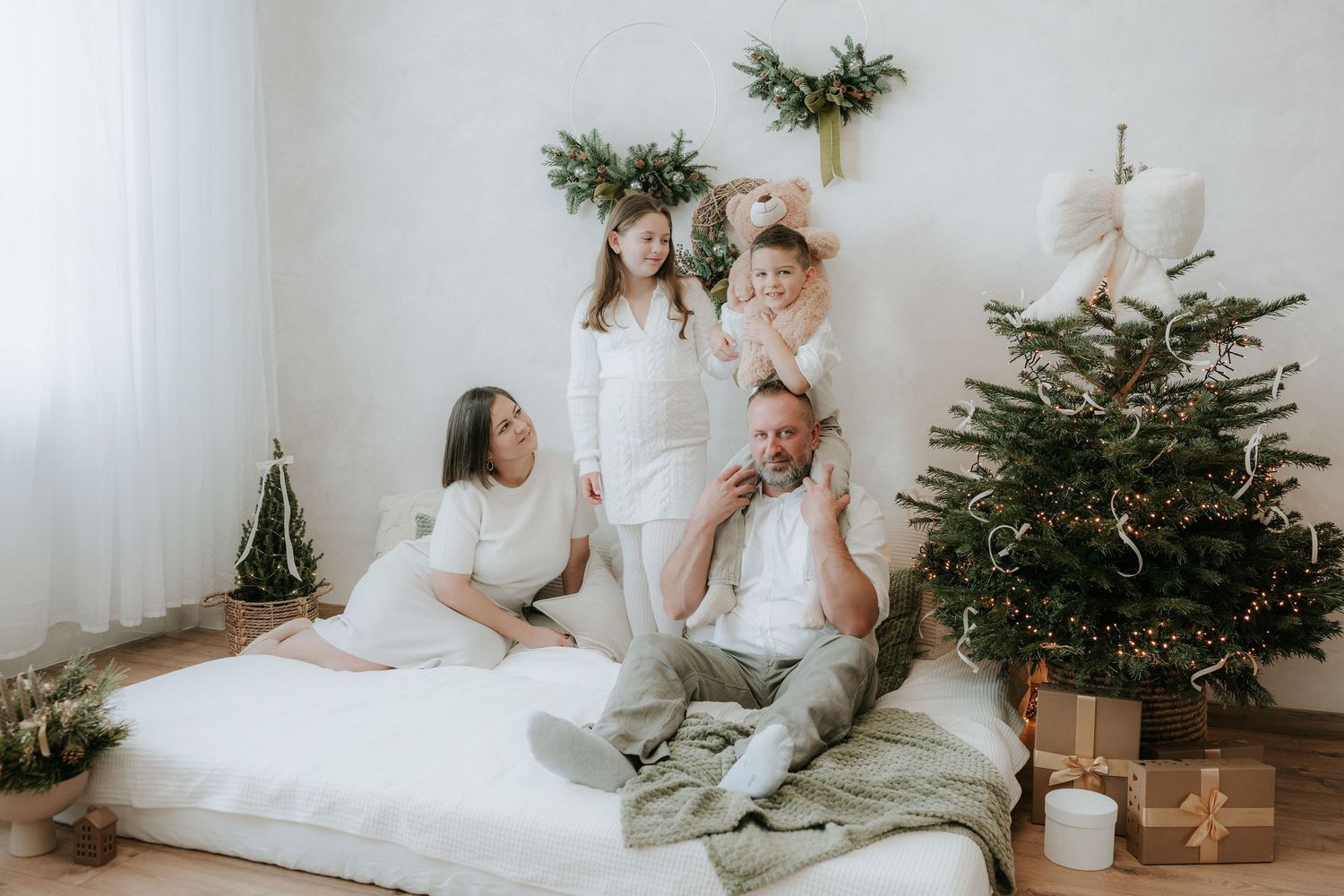 Family in white outfits celebrating Christmas near a decorated tree with gifts in a bright, cozy room.