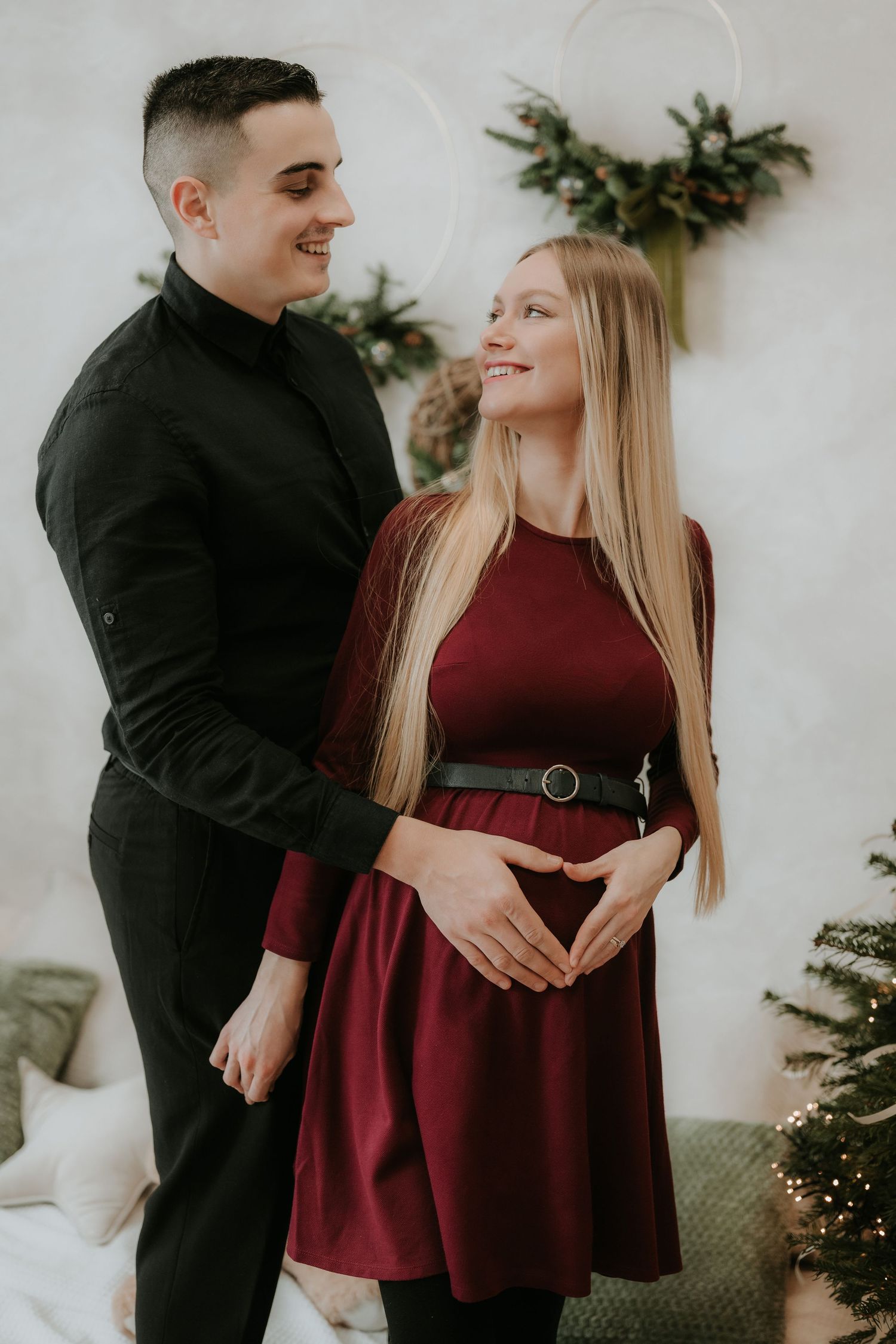 Smiling couple in festive holiday setting, man in black embracing pregnant woman in burgundy dress near Christmas tree.