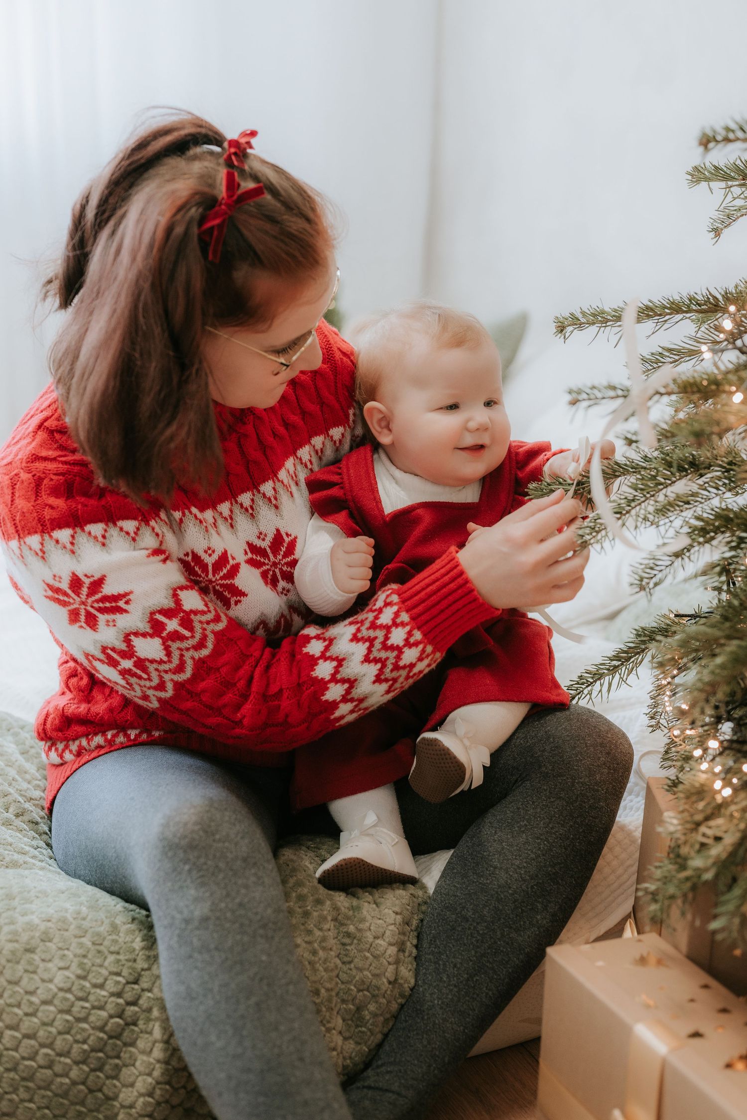 Mother and baby in matching red Nordic sweaters decorating a Christmas tree together on a cozy bed.