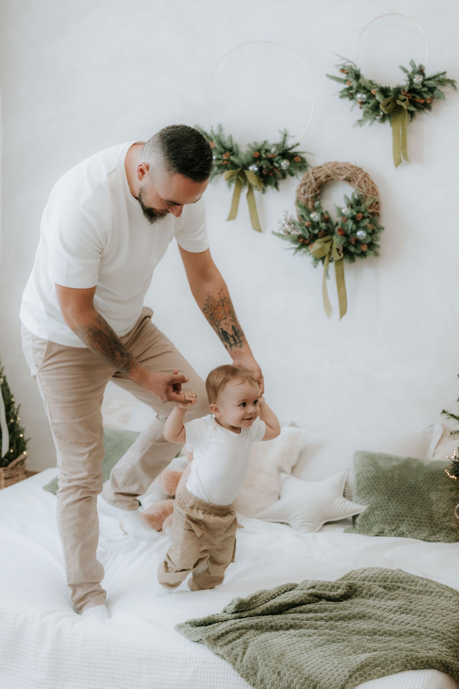 Father helping baby stand on bed with Christmas wreaths on white wall in festive holiday home setting.