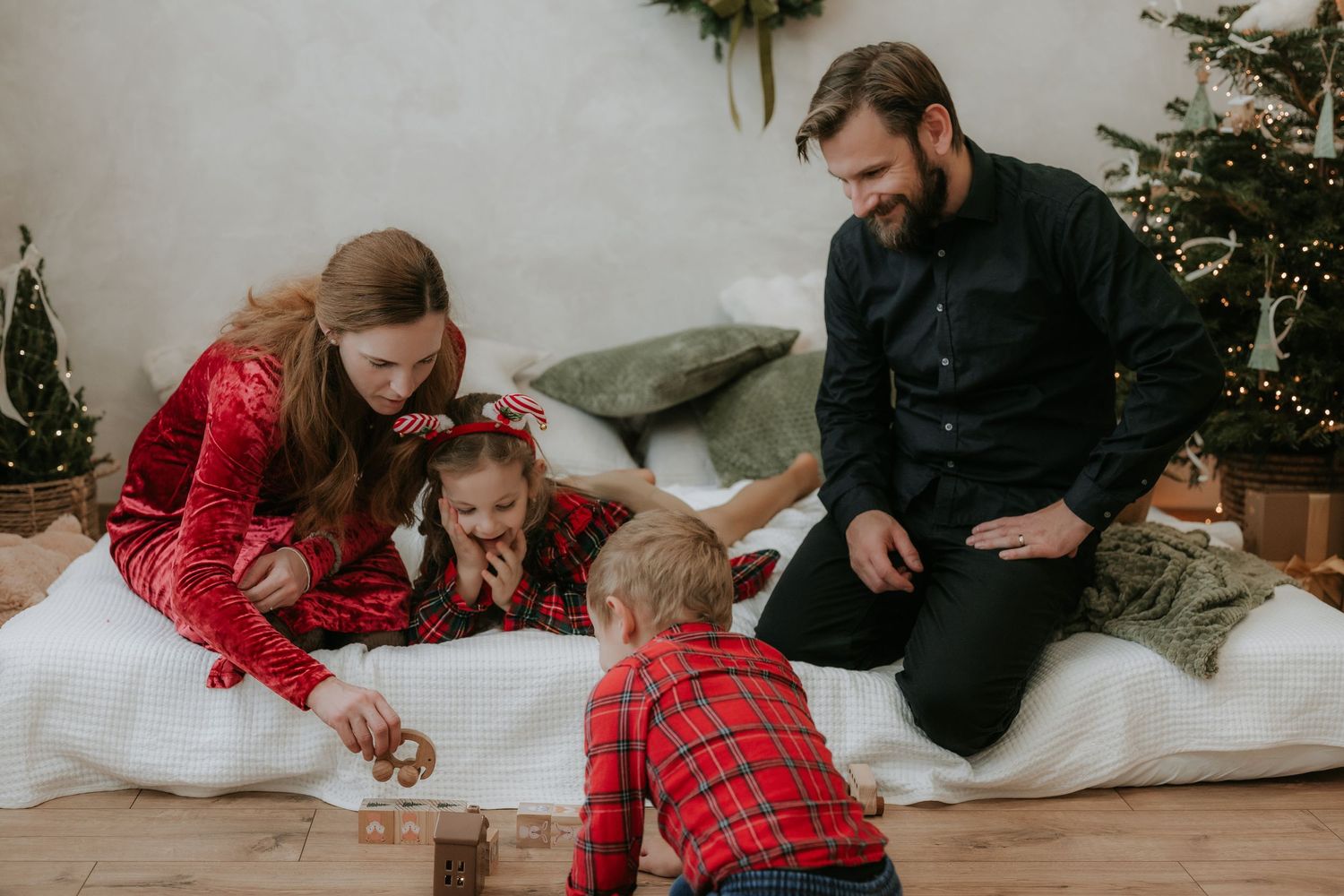 Family in red and black holiday pajamas playing with wooden blocks near a decorated Christmas tree on a white blanket.