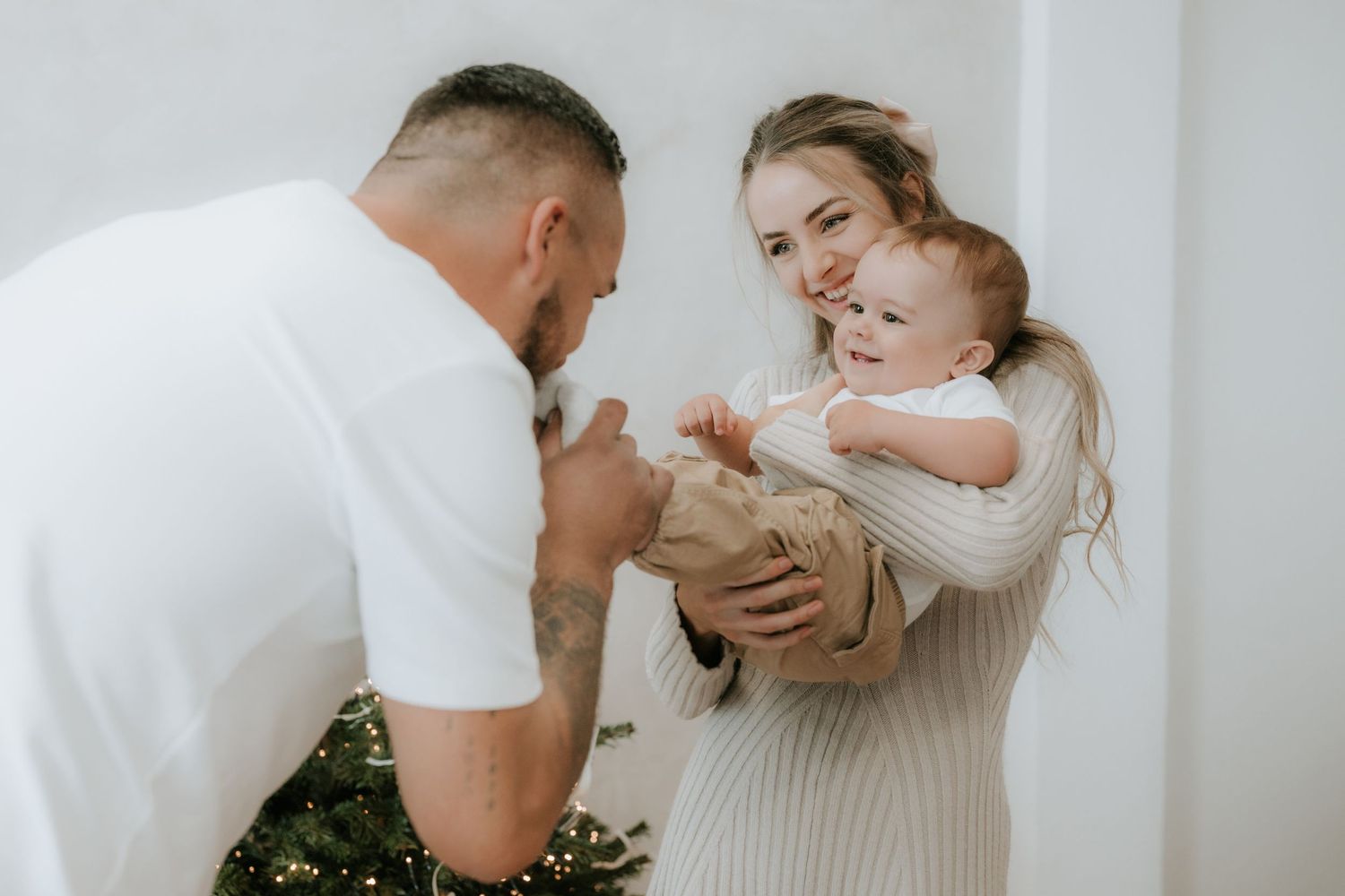 A young family with tattoos shares a joyful moment, as a mother holds her baby while a father interacts lovingly indoors.