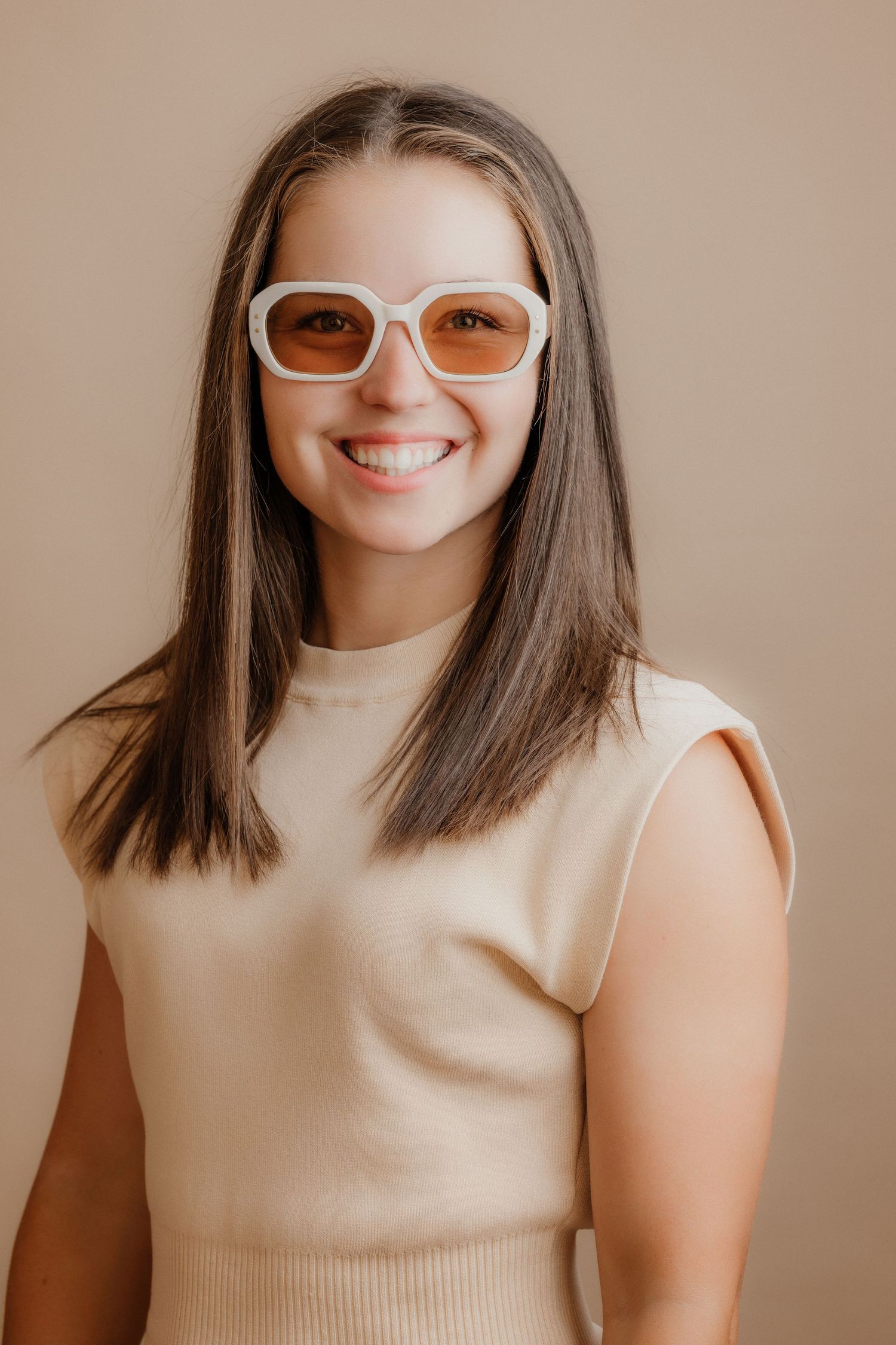Smiling brunette teenage girl wearing white sunglasses and beige sleeveless top on neutral background.
