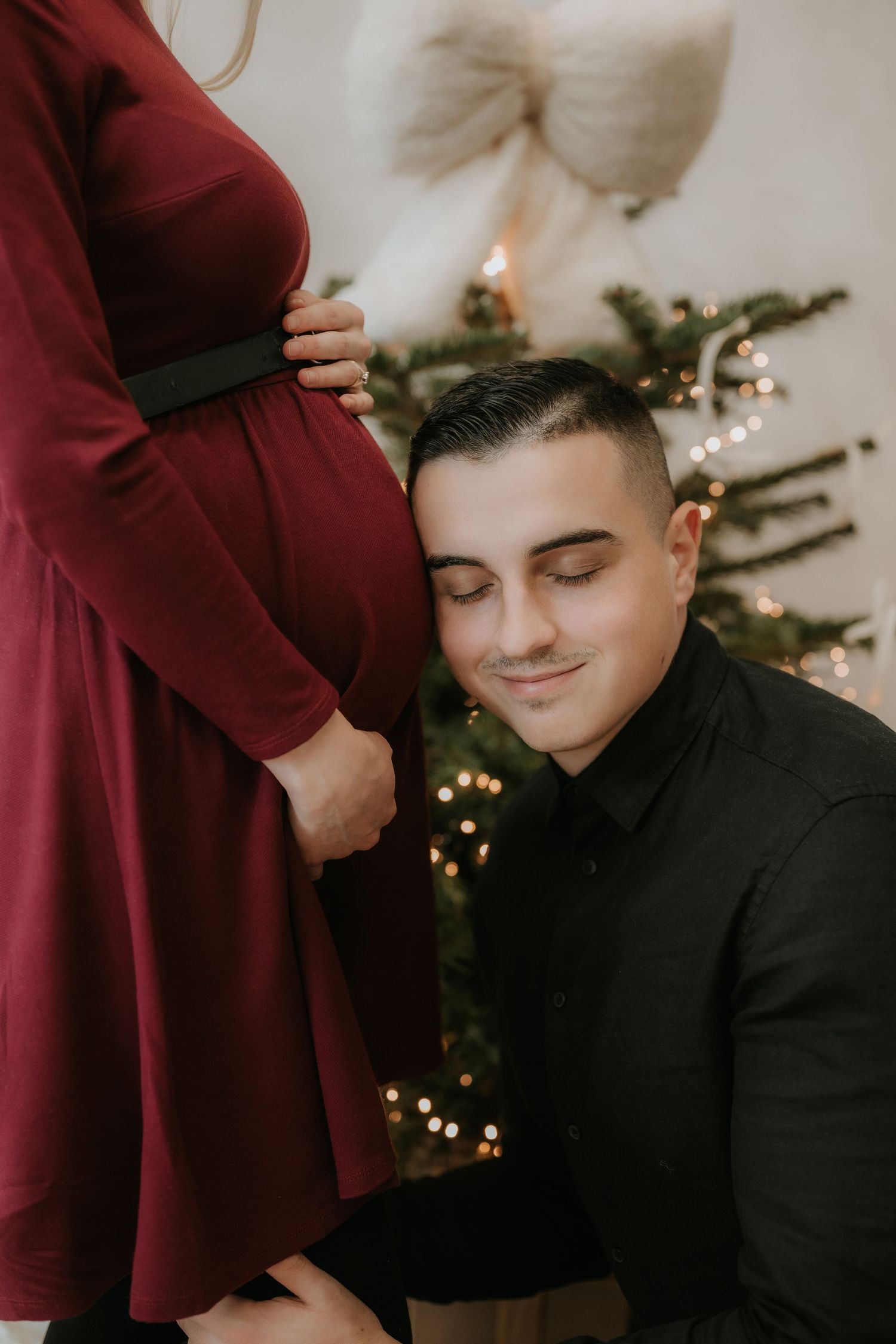 Man kneeling beside pregnant woman in red dress near Christmas tree with bokeh lights.