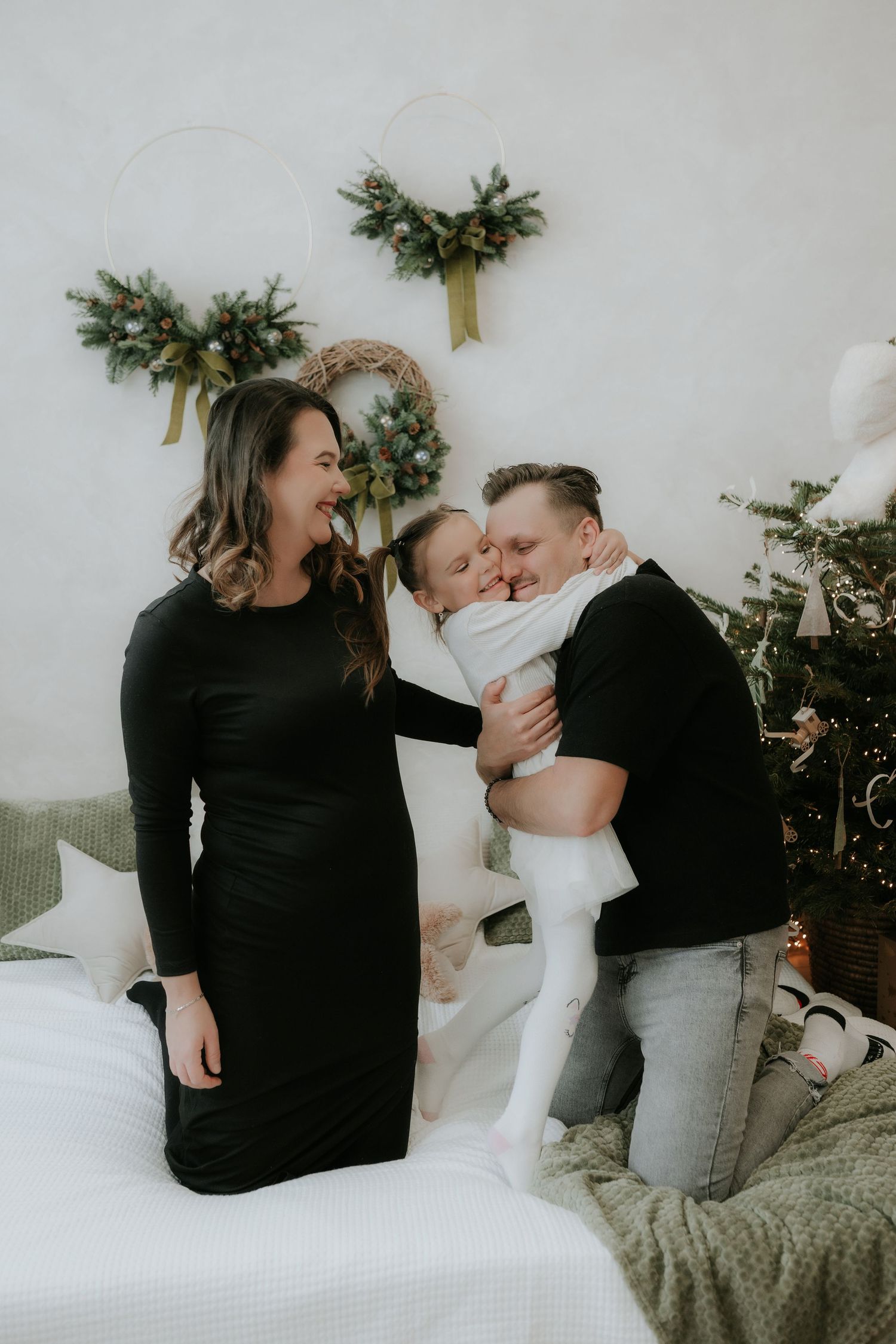 Family in black outfits celebrating Christmas, father kissing toddler while mother smiles near decorated tree.