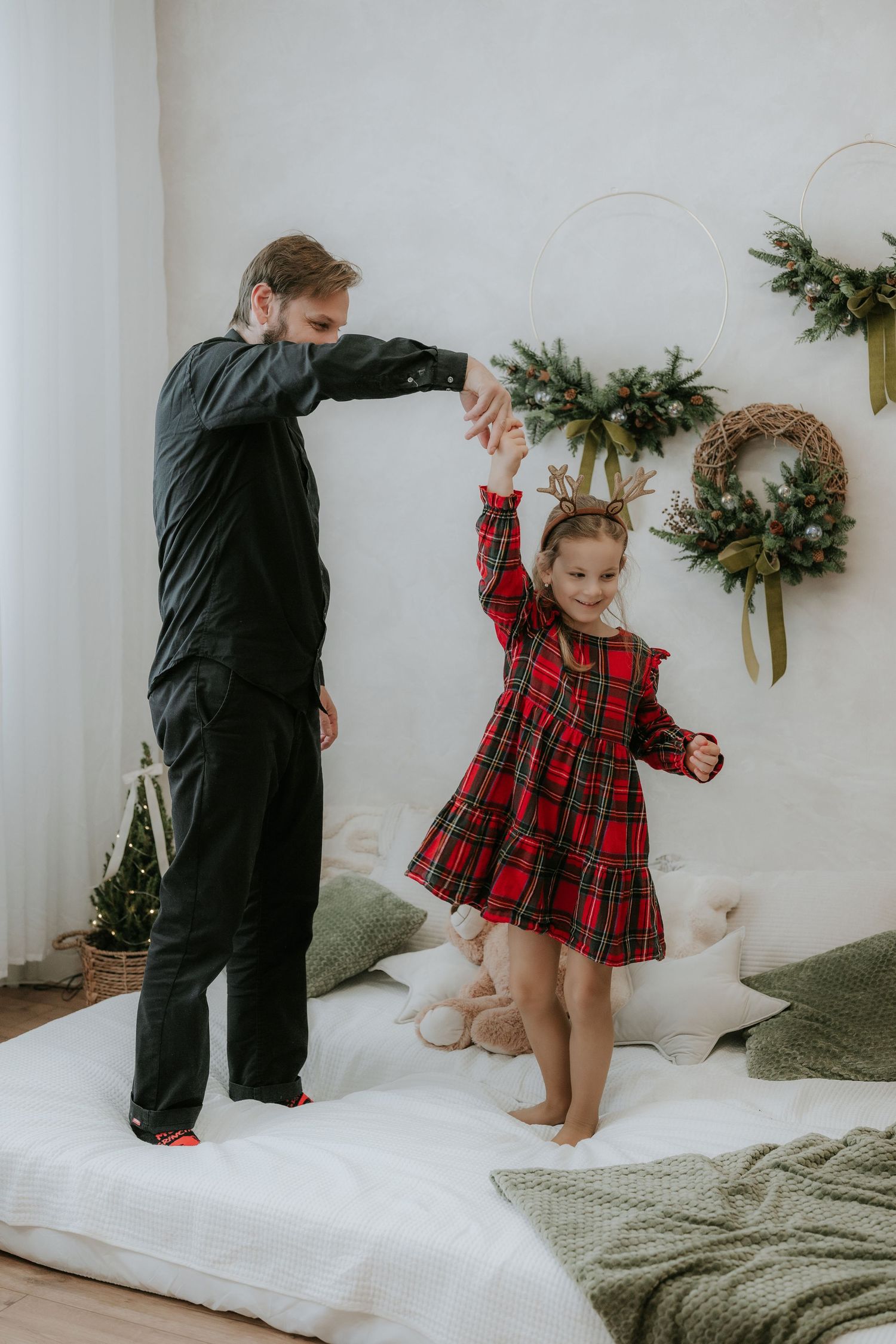 Father and daughter dancing on bed in Christmas decorated room, girl wearing red plaid dress with reindeer antlers headband.