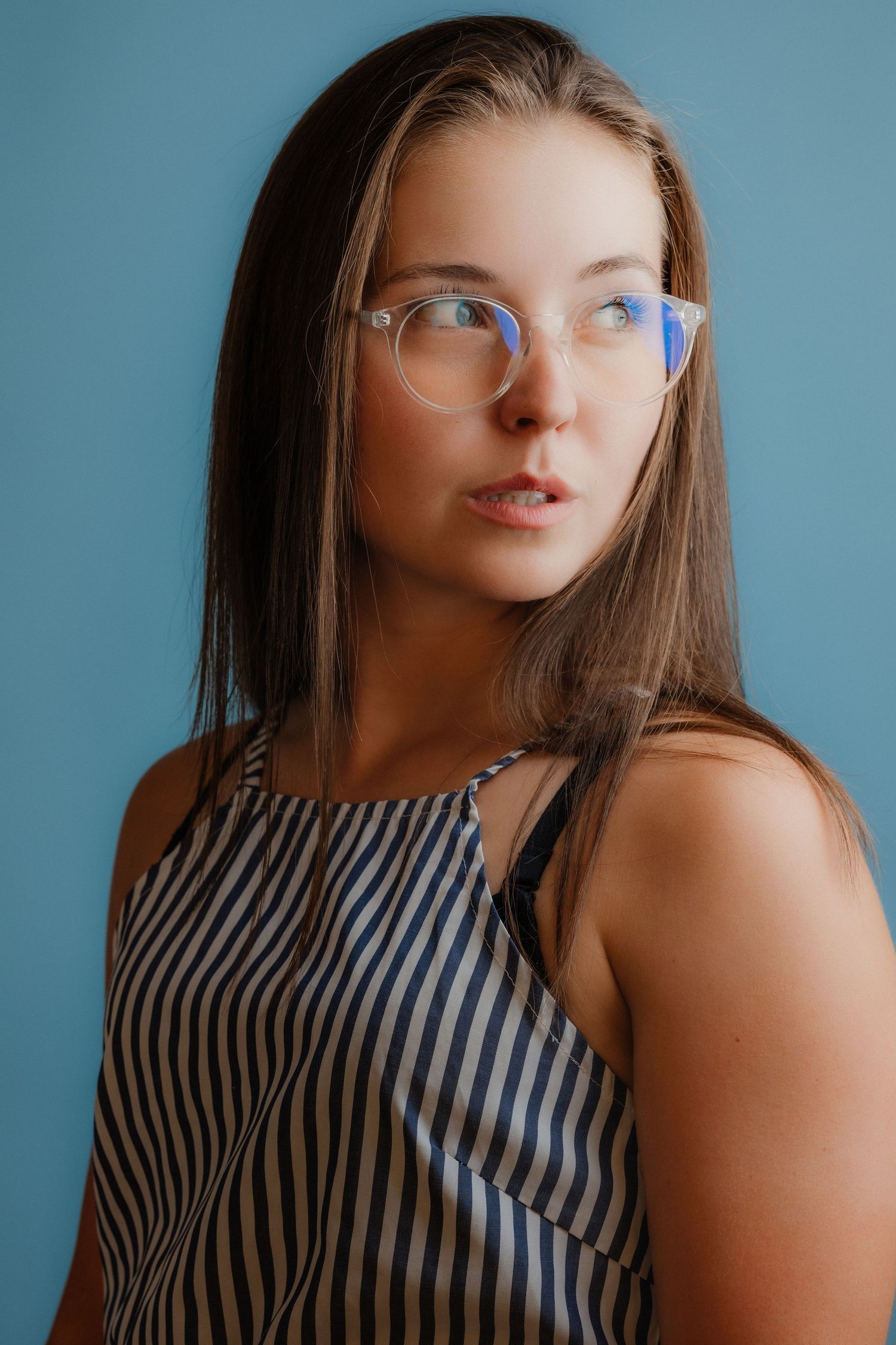 Young woman with long brown hair and round glasses wearing a striped top, posing against a blue background.
