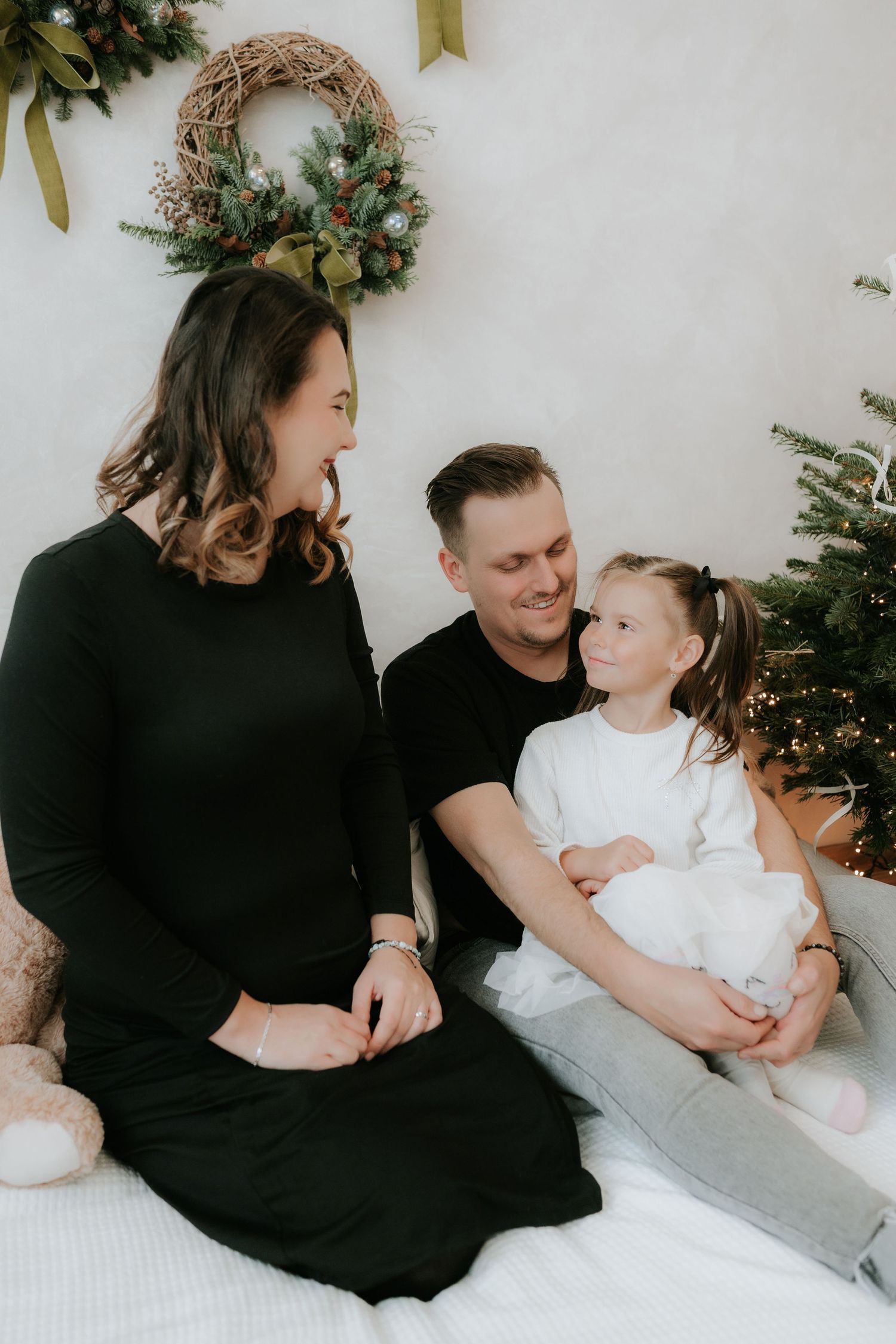 Family of three sharing a joyful moment by a decorated Christmas tree with festive wreath backdrop.