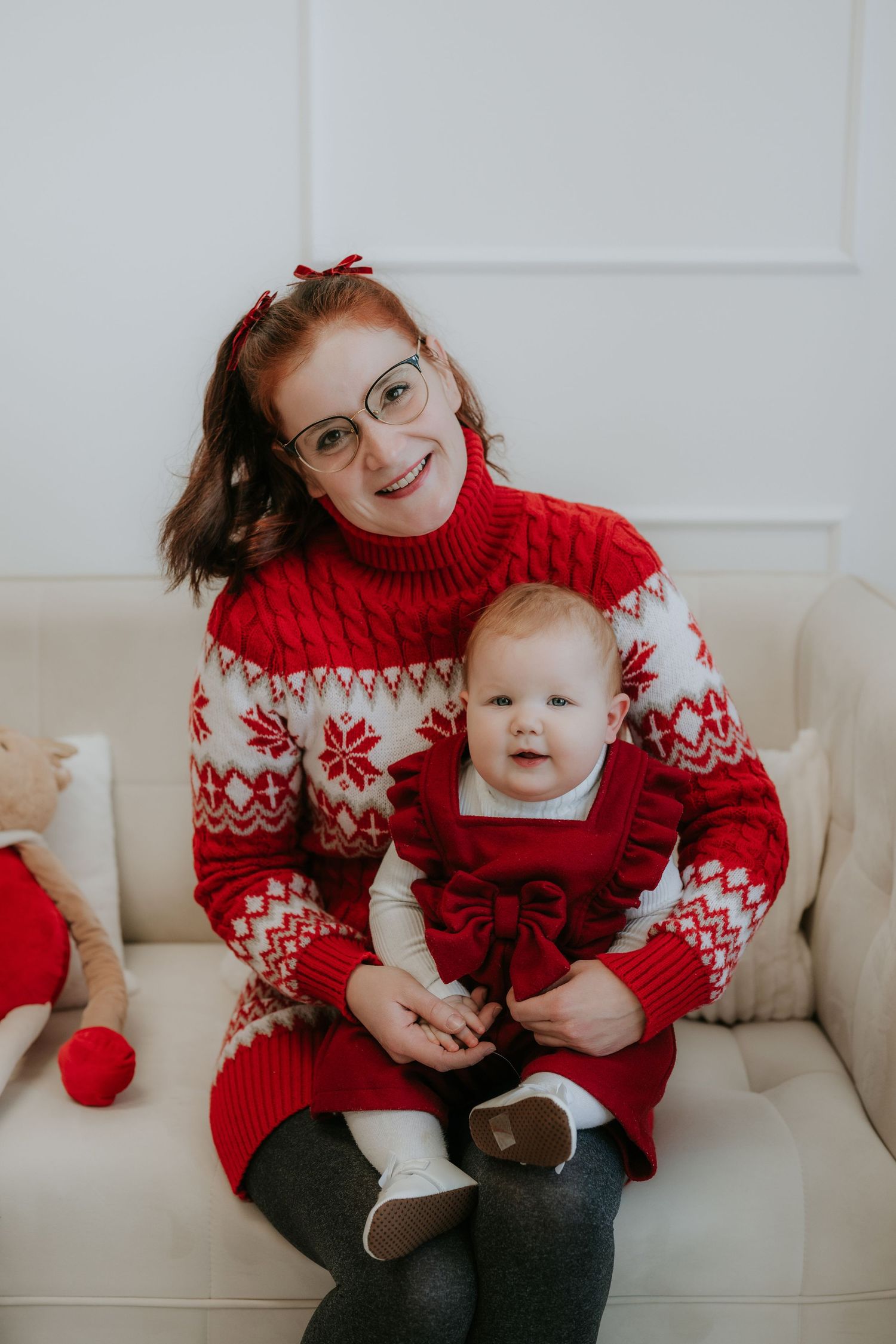 A smiling redhead woman in a red Nordic sweater holds a baby girl in a matching red dress on a white sofa.