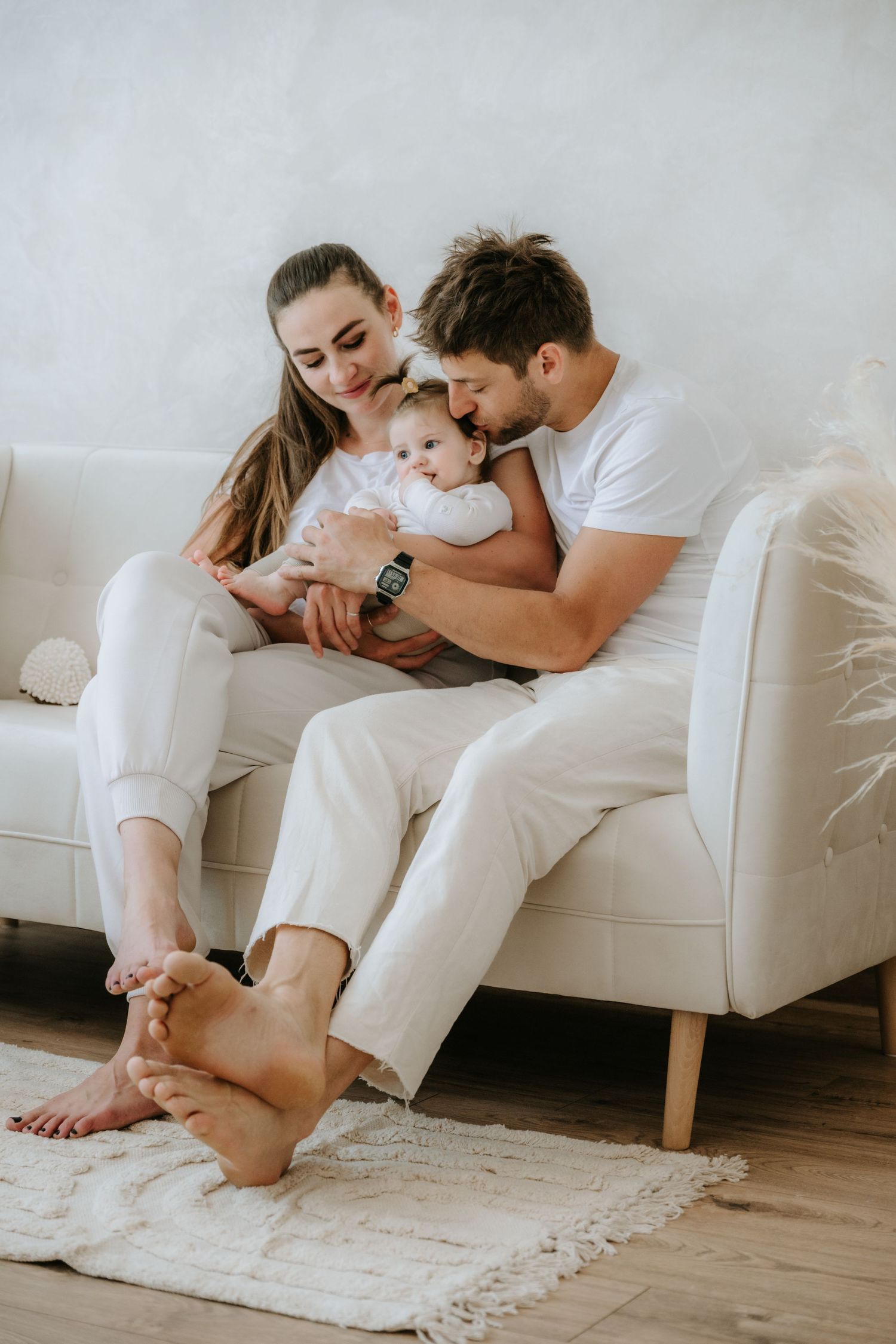Young family with baby relaxing on white sofa, parents in white outfits sharing tender moment with infant.