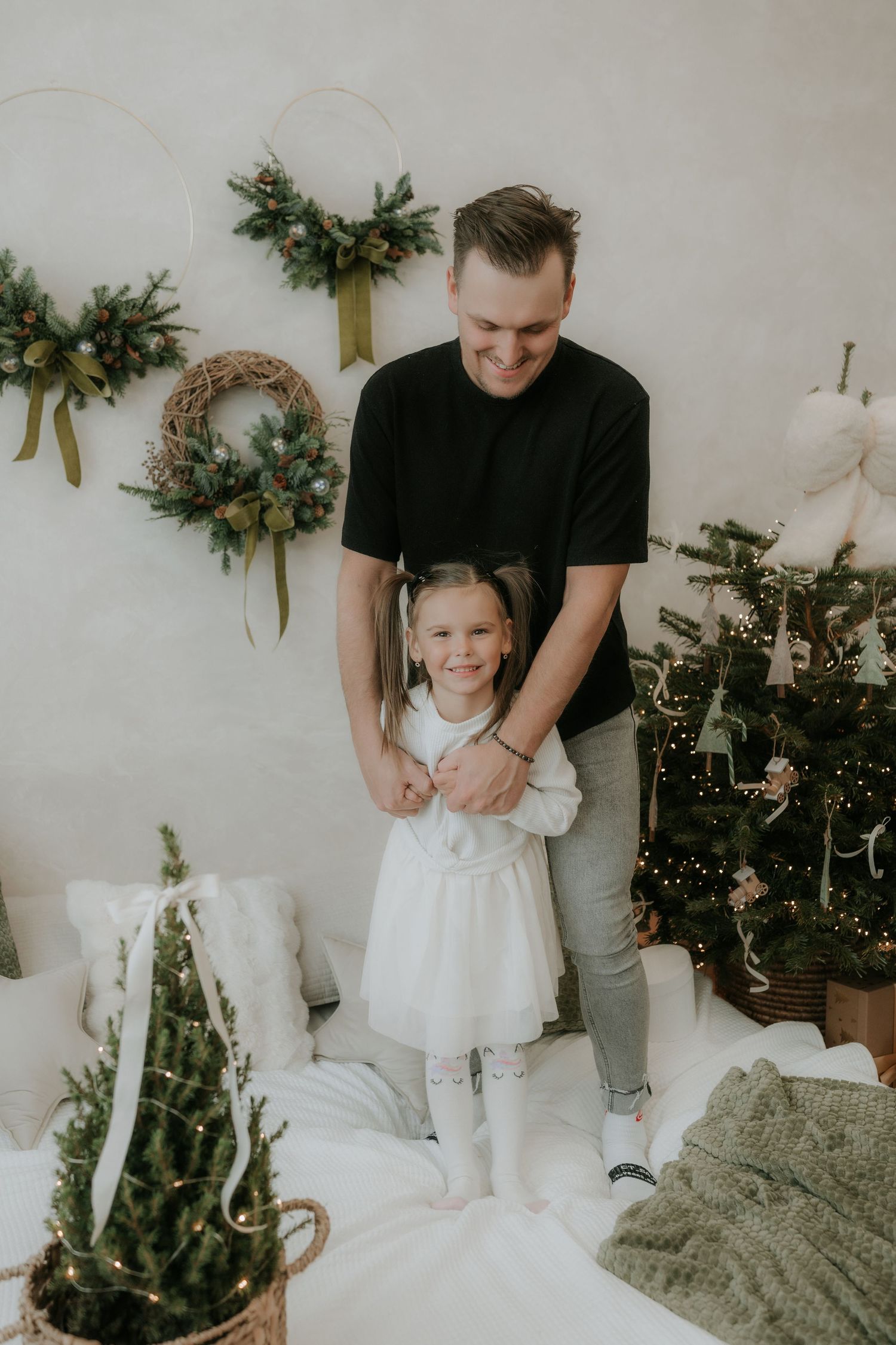 Father smiling and holding young daughter in white dress near decorated Christmas tree and holiday wreaths.