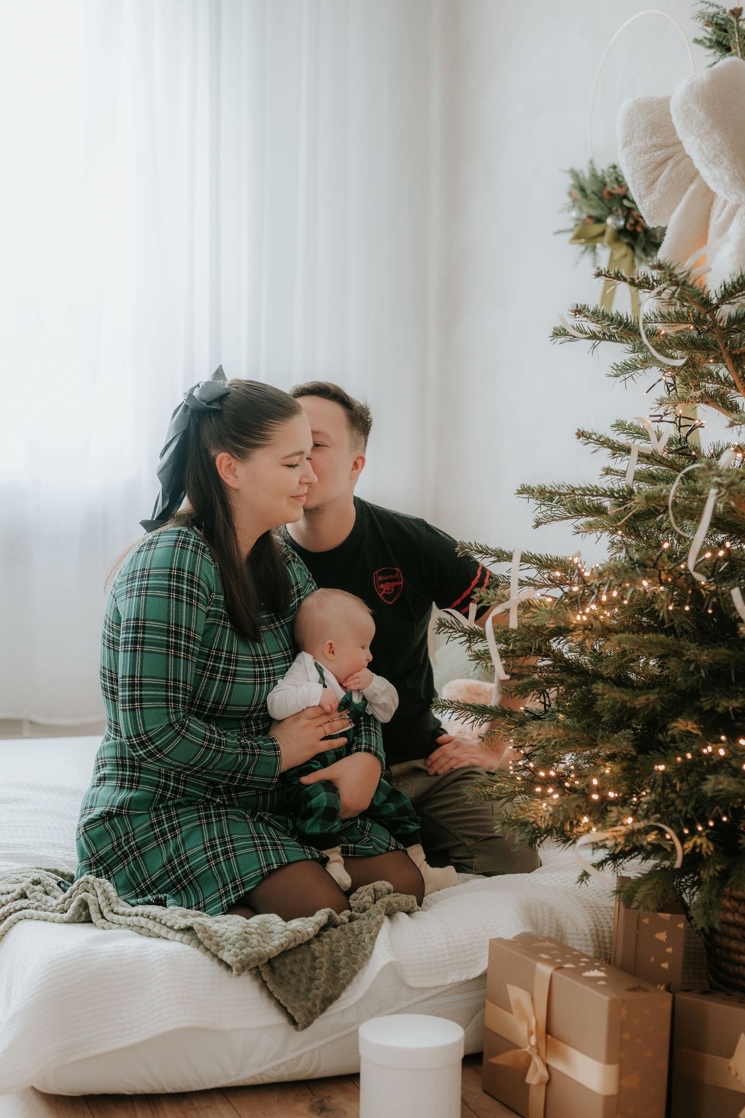 A young family sits together by a decorated Christmas tree, with mother holding a baby while father looks on lovingly.