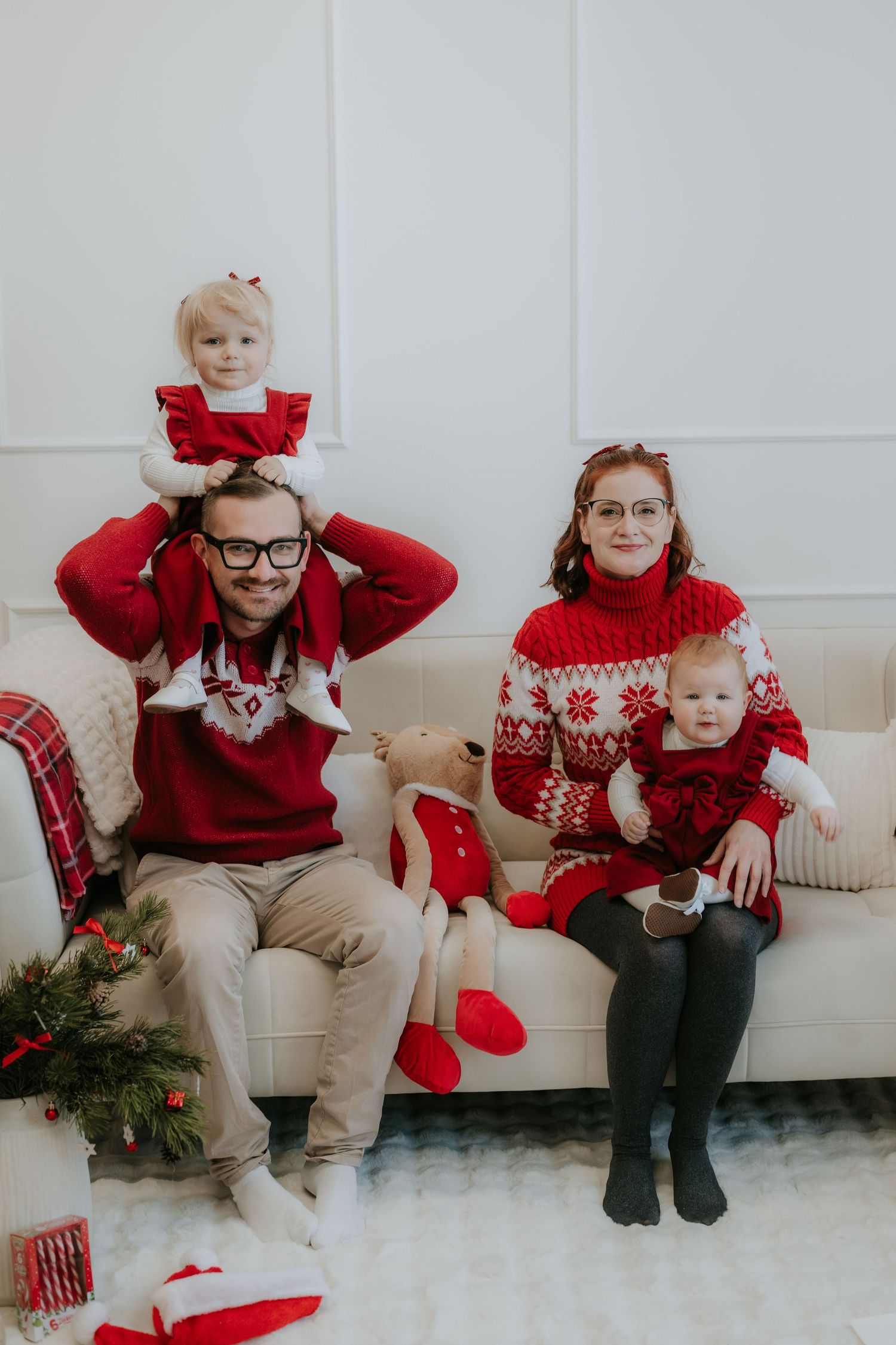 Family in matching red Christmas sweaters poses on white sofa with two young children during holiday photo session.
