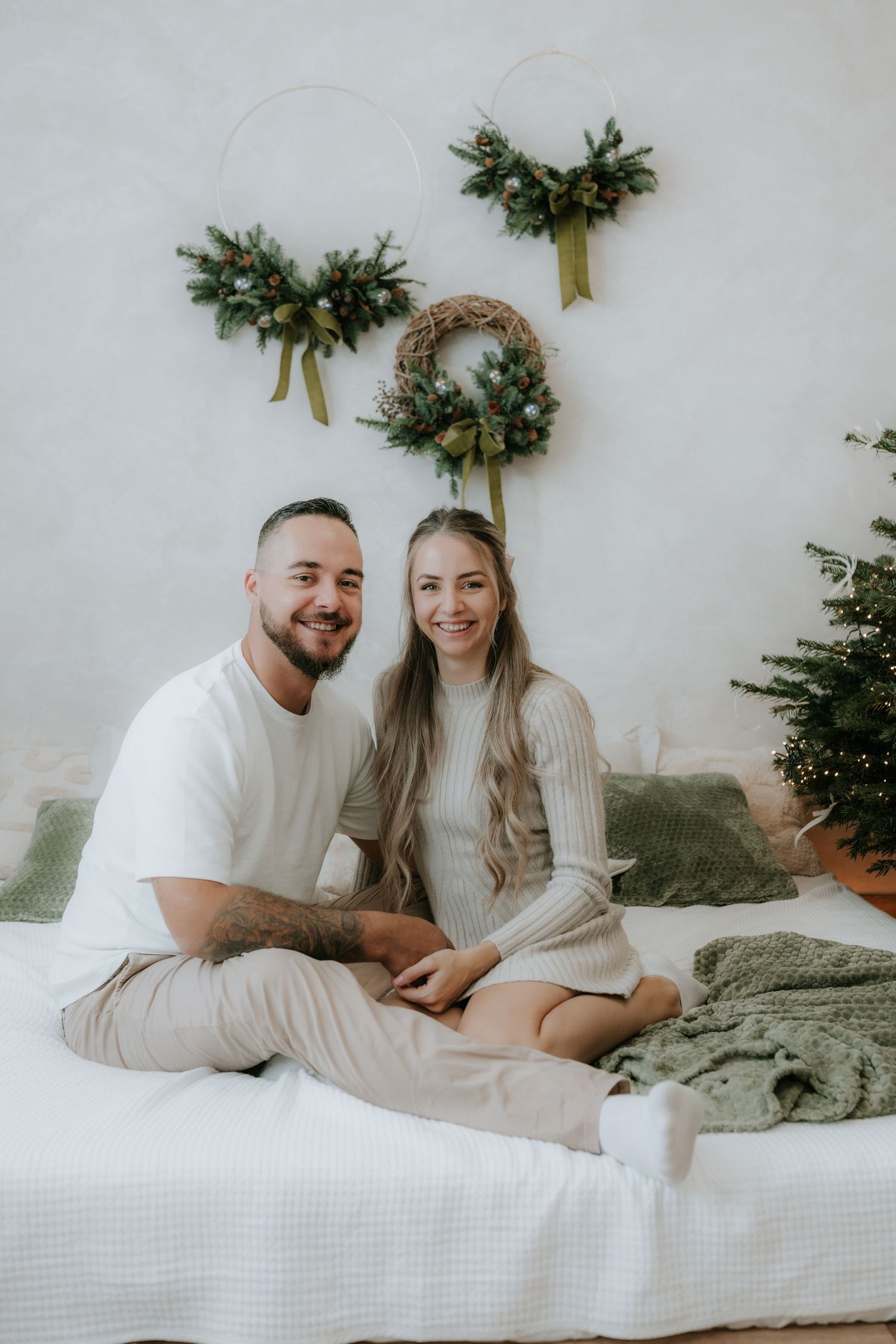Happy couple sitting on a bed with Christmas wreaths on the wall and a decorated tree nearby.