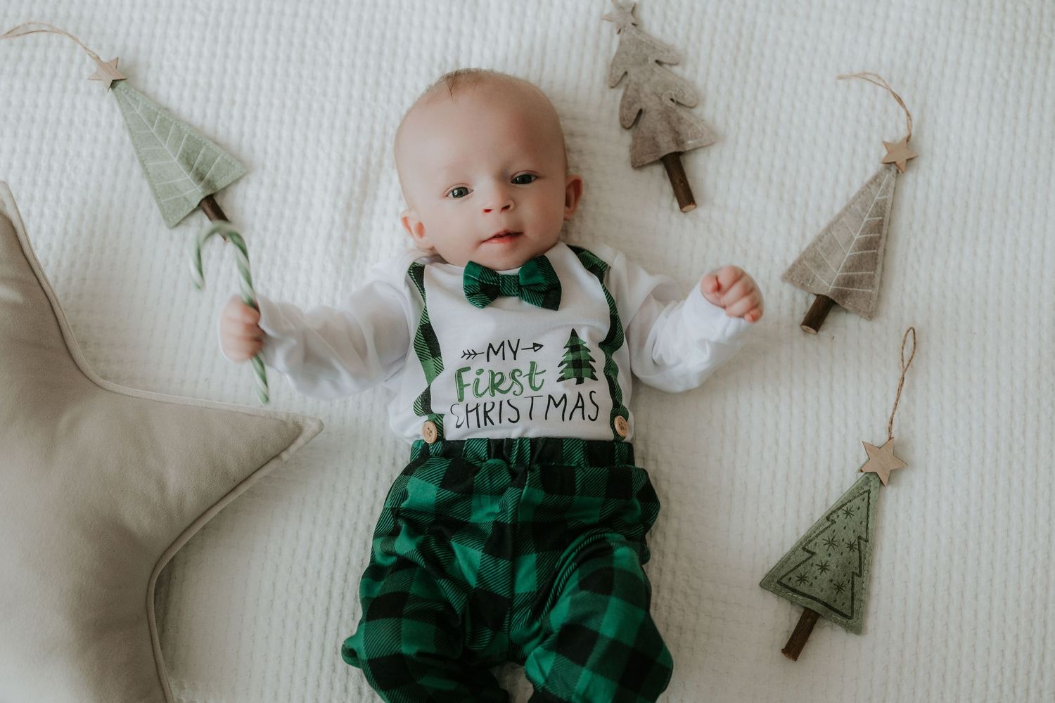 Baby in green plaid pants and bow tie bib reading My First Christmas, surrounded by Christmas tree ornaments.