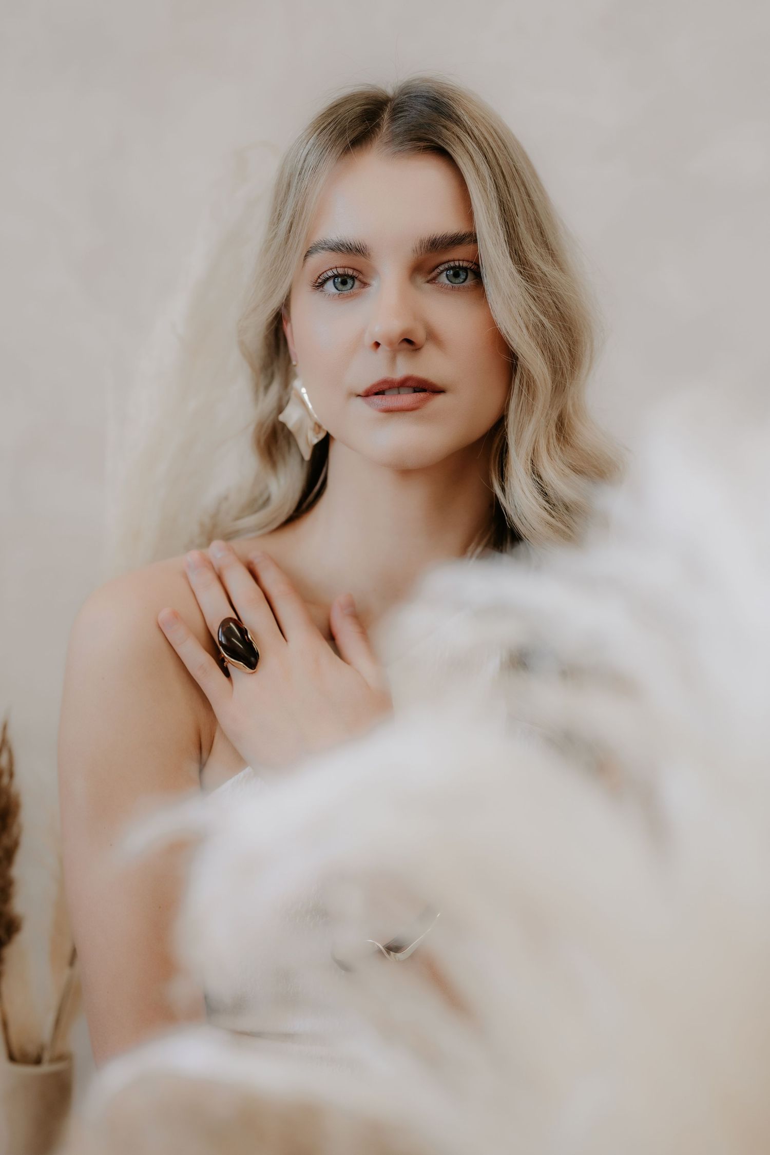 Blonde woman with blue eyes wearing a dark ring, posing gracefully against a neutral beige background with soft white florals.