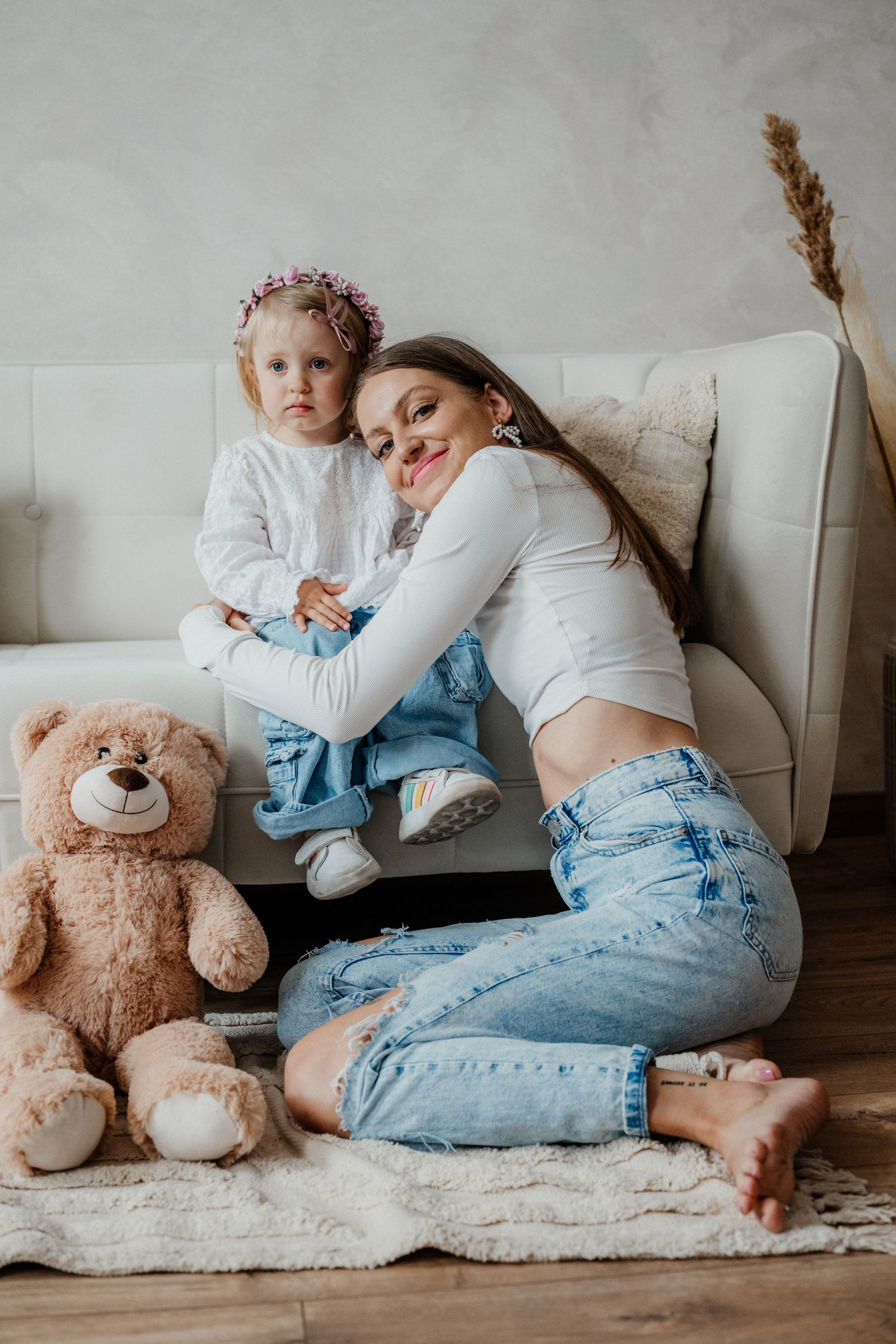 Young mother in white top and jeans sits on floor hugging toddler daughter on sofa with teddy bear nearby.