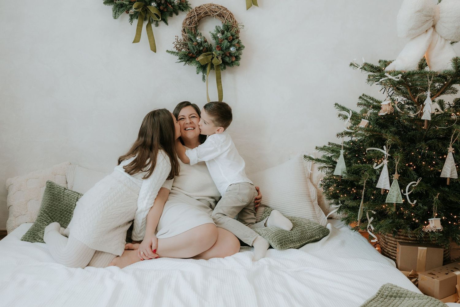 Family in white outfits celebrating Christmas near decorated tree, mother receiving kisses from children on white bed.