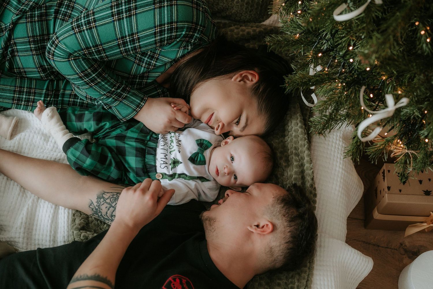 Family in green plaid pajamas lying by a Christmas tree, mother kissing baby while toddler snuggles close.