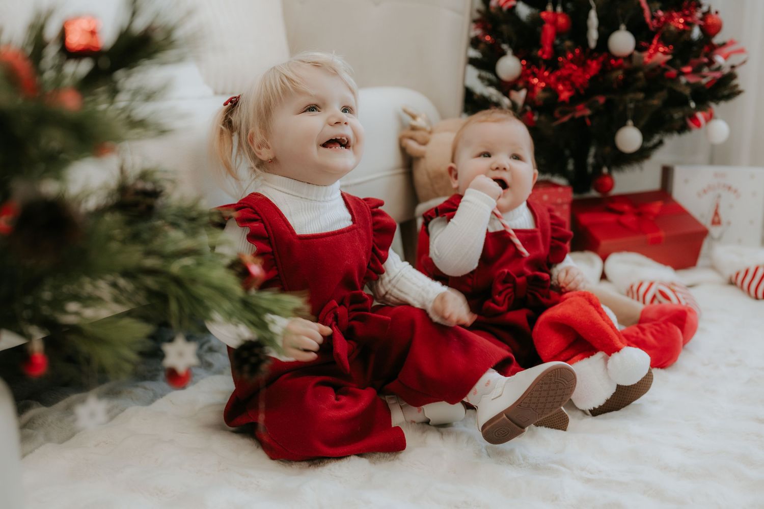 Two toddlers in matching red Christmas dresses sitting together near a decorated holiday tree.