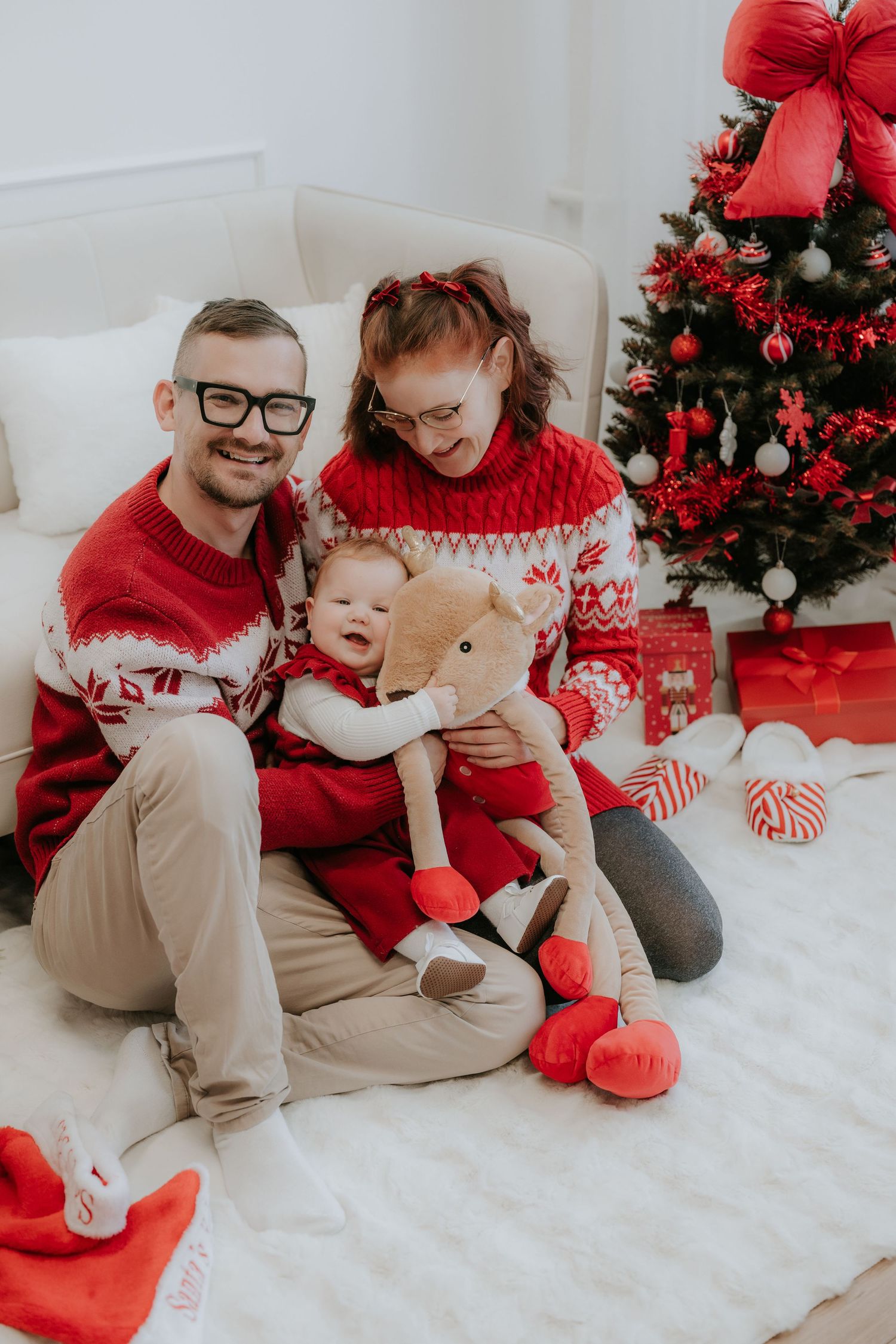 Happy family in matching red Christmas sweaters sitting by decorated tree with baby holding teddy bear.