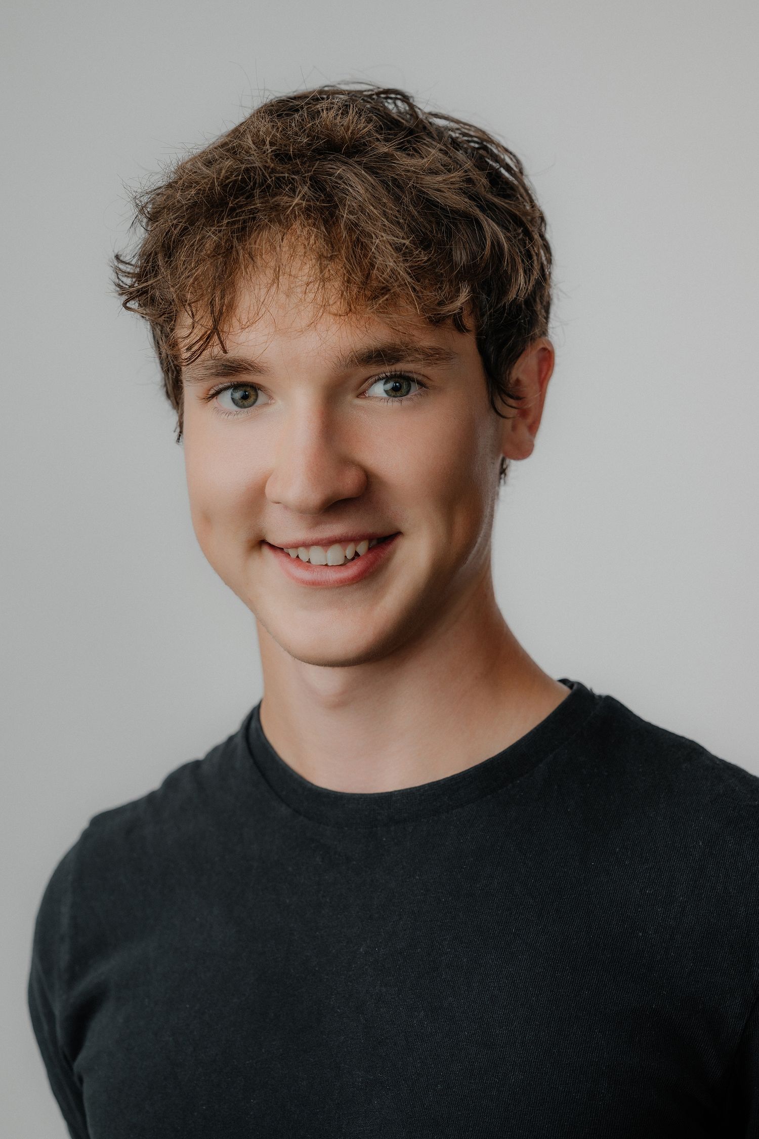 Young man with curly brown hair and blue eyes smiling, wearing a dark green shirt against a gray background.