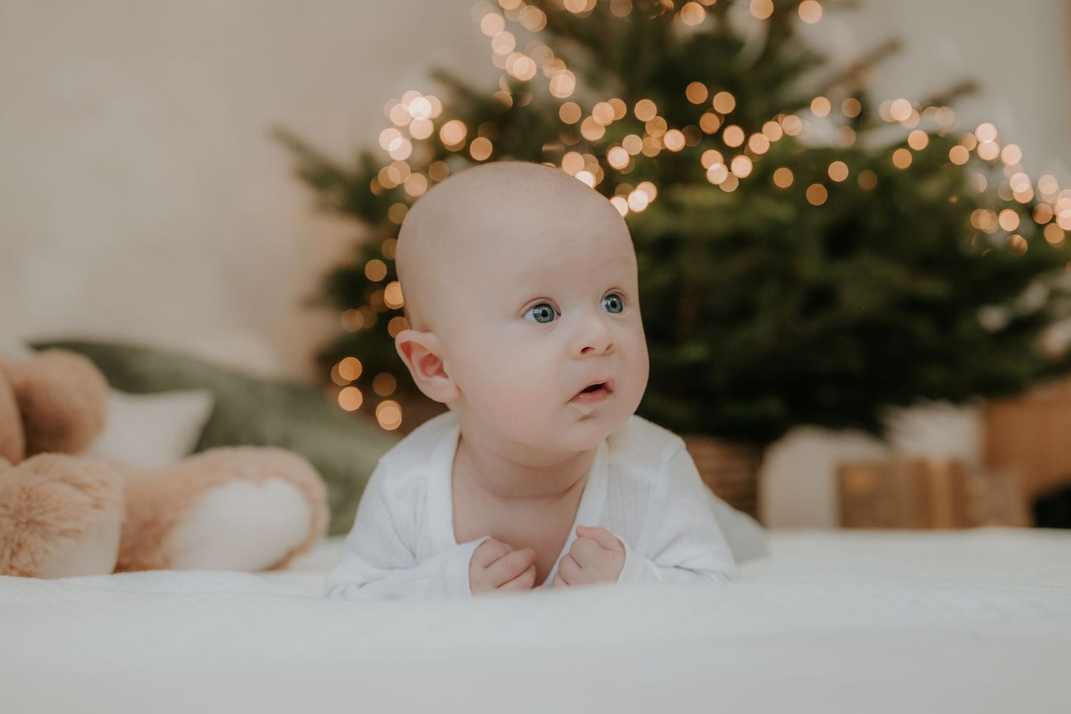 A curious baby in a white onesie does tummy time near a decorated Christmas tree with warm bokeh lights.
