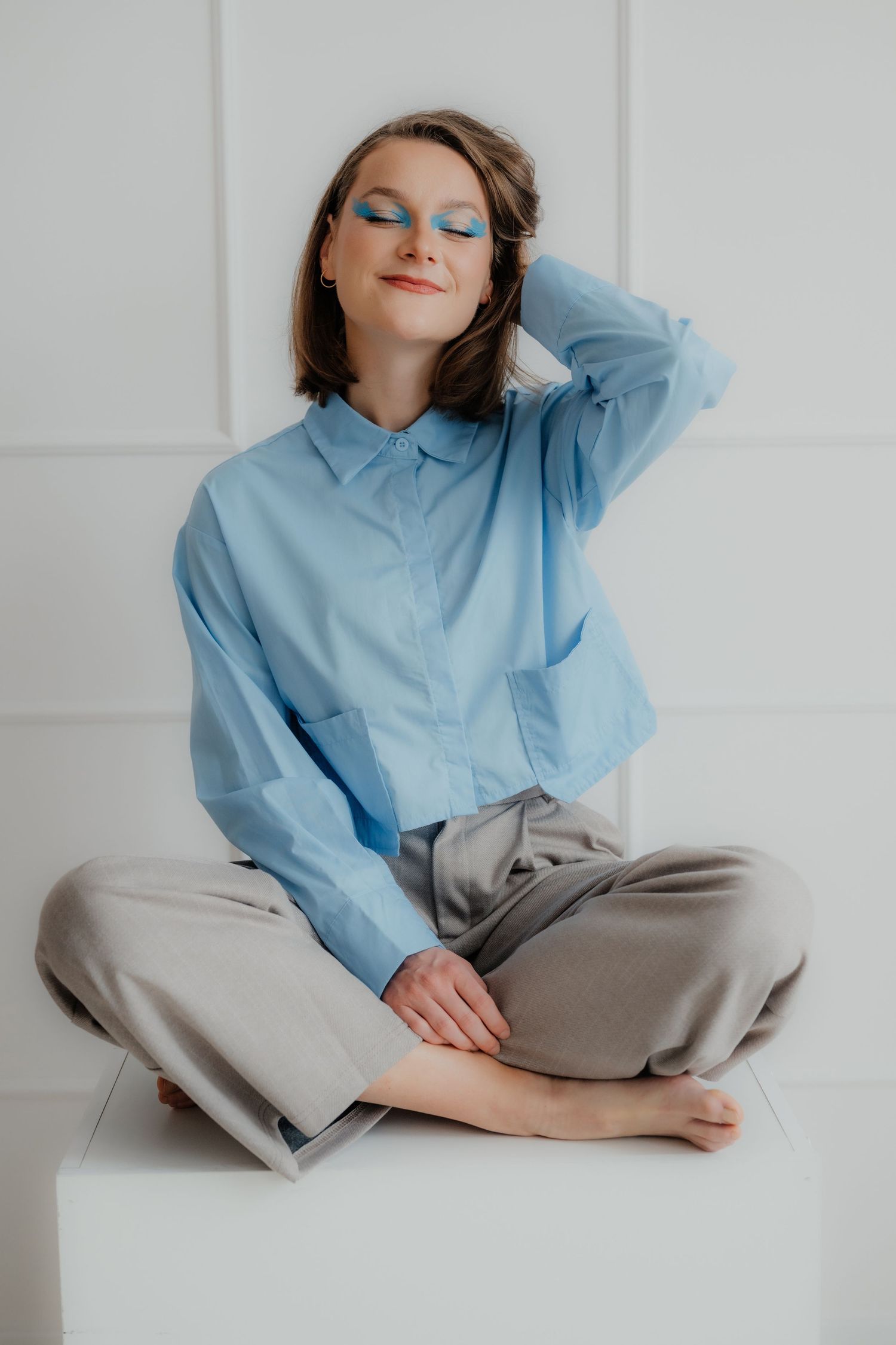 Woman in blue shirt and gray pants sitting cross-legged on white cube with bold blue eye makeup.