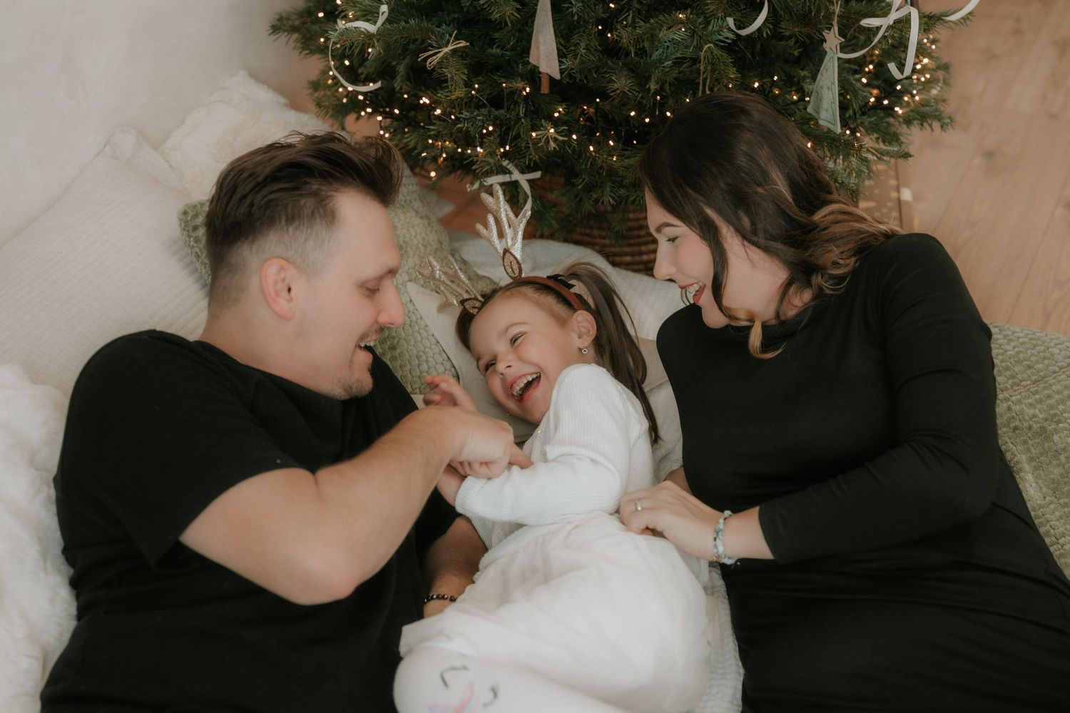 Family laughing together lying by a decorated Christmas tree, wearing black and white outfits in a cozy holiday setting.