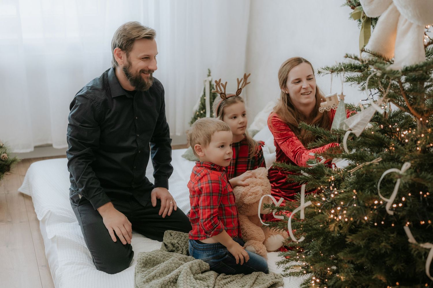 Family decorating a Christmas tree together in a bright white living room, parents and two young children in festive red pajamas.