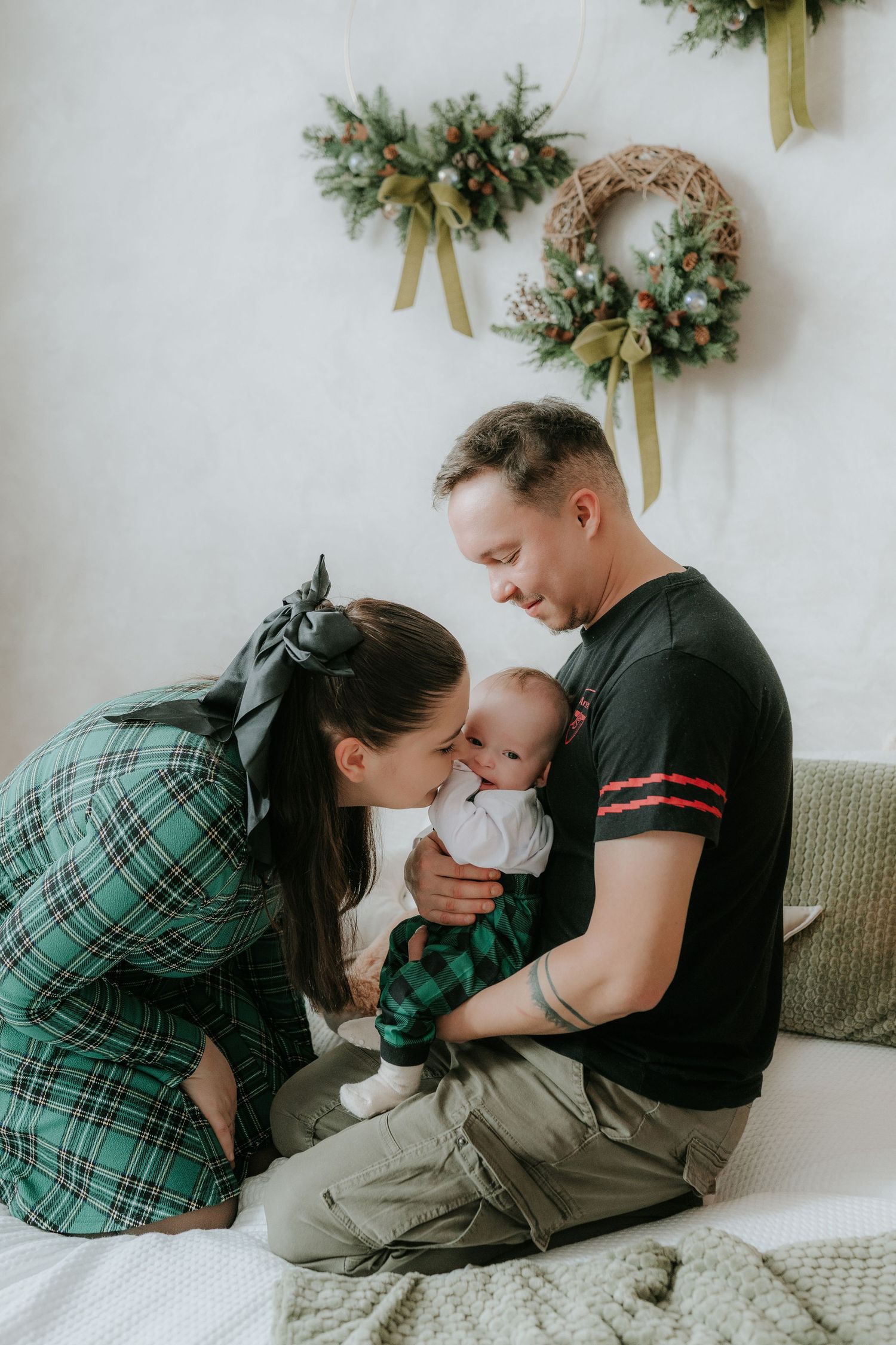 Young family with newborn baby celebrating Christmas, parents kissing infant near holiday wreaths on white wall.